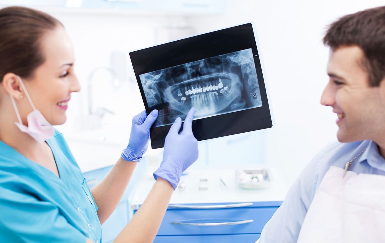 Dentist in blue scrubs points to dental X-ray, smiling at a patient in a dental chair.
