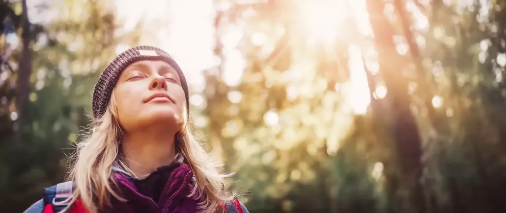 A person in a beanie and outdoor gear stands in a sunlit forest with eyes closed, breathing deeply.
