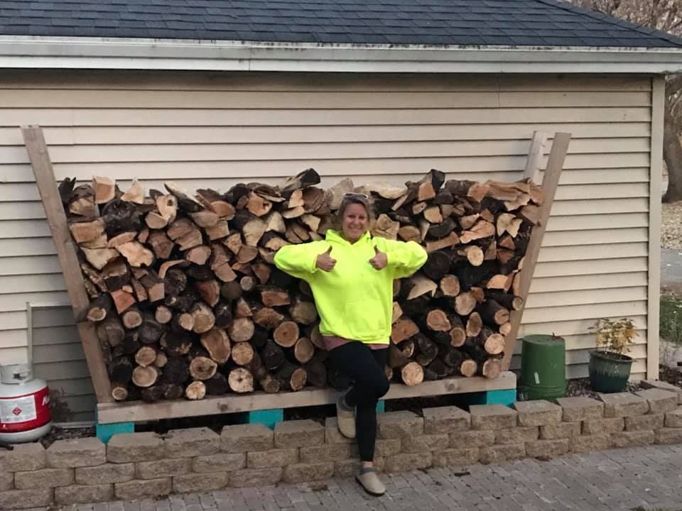 A woman in a neon yellow sweater is standing in front of a pile of logs.