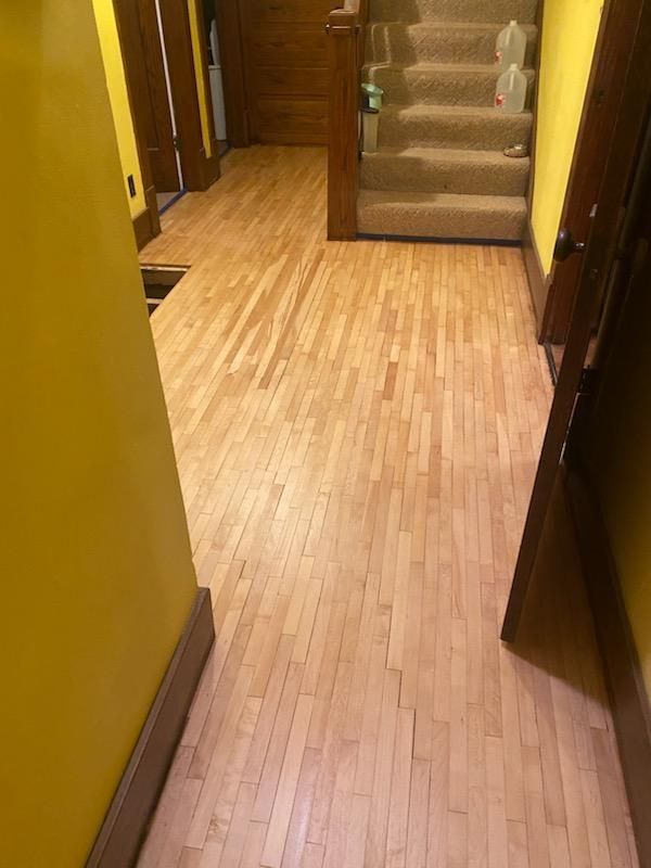 A hallway with a wooden floor and stairs in the background.