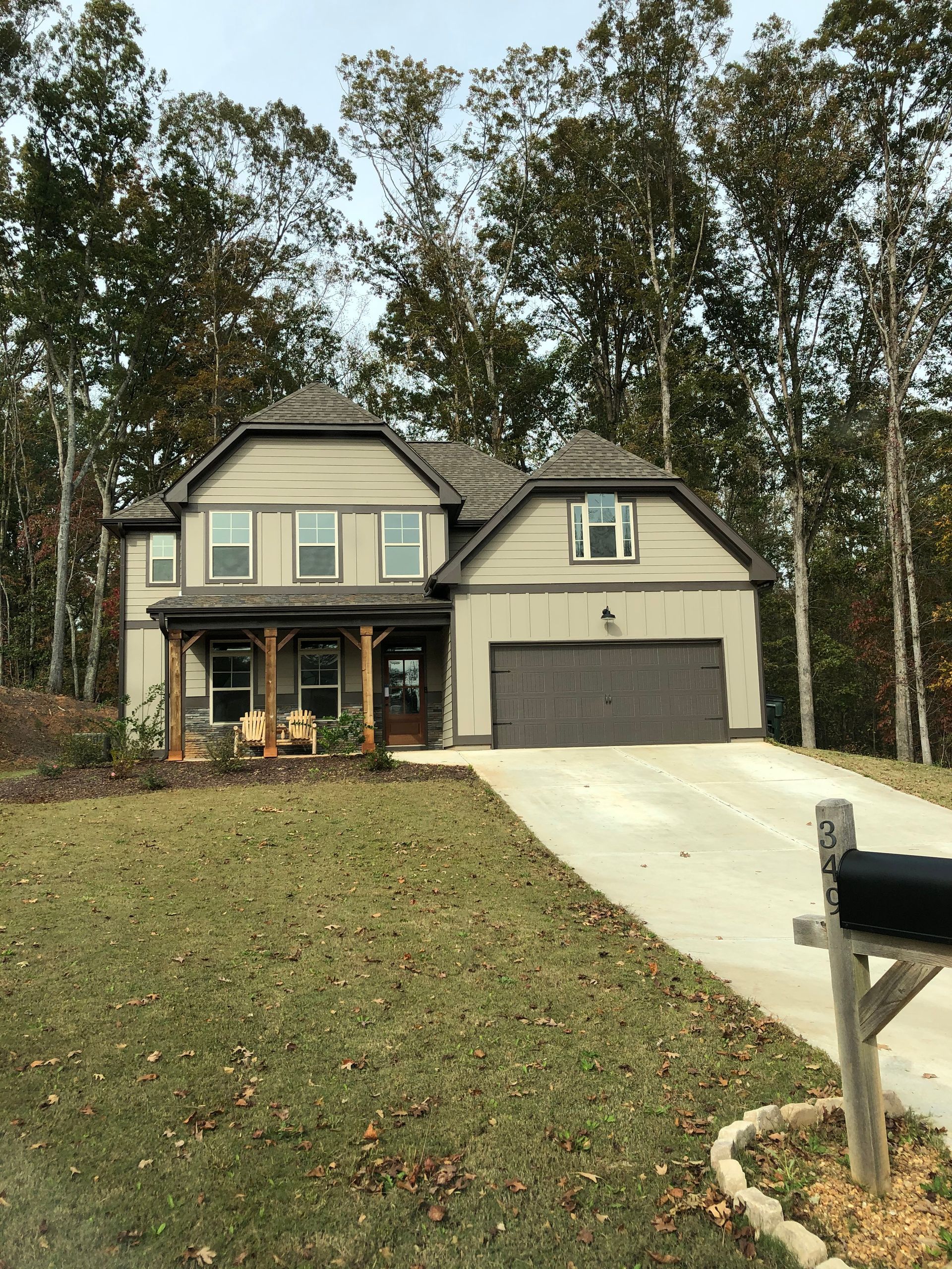 Two-story beige house with a brown garage door and porch, set in a wooded area.