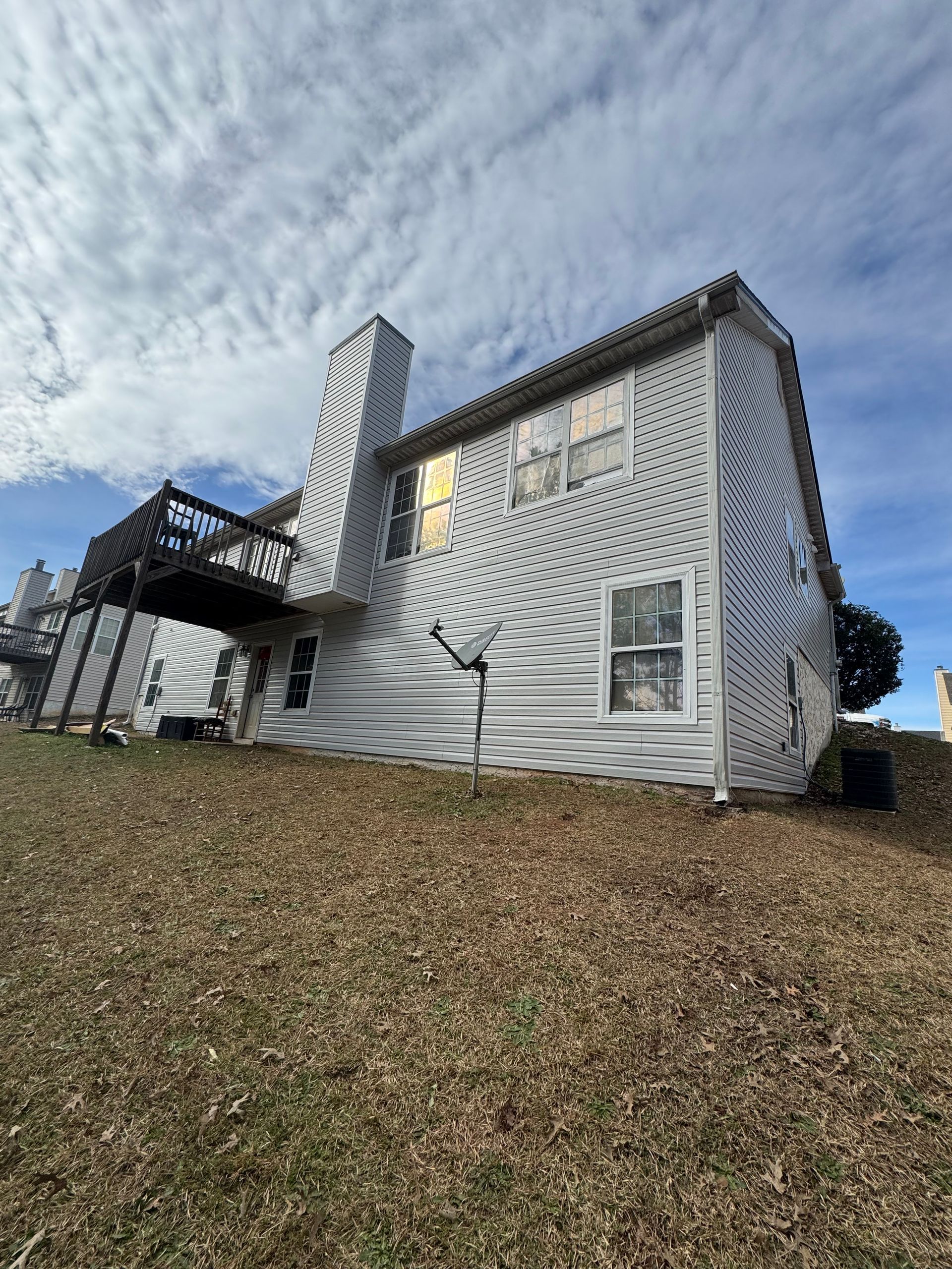 Two-story house with white siding, chimney, and deck on a grassy hill under a cloudy sky.
