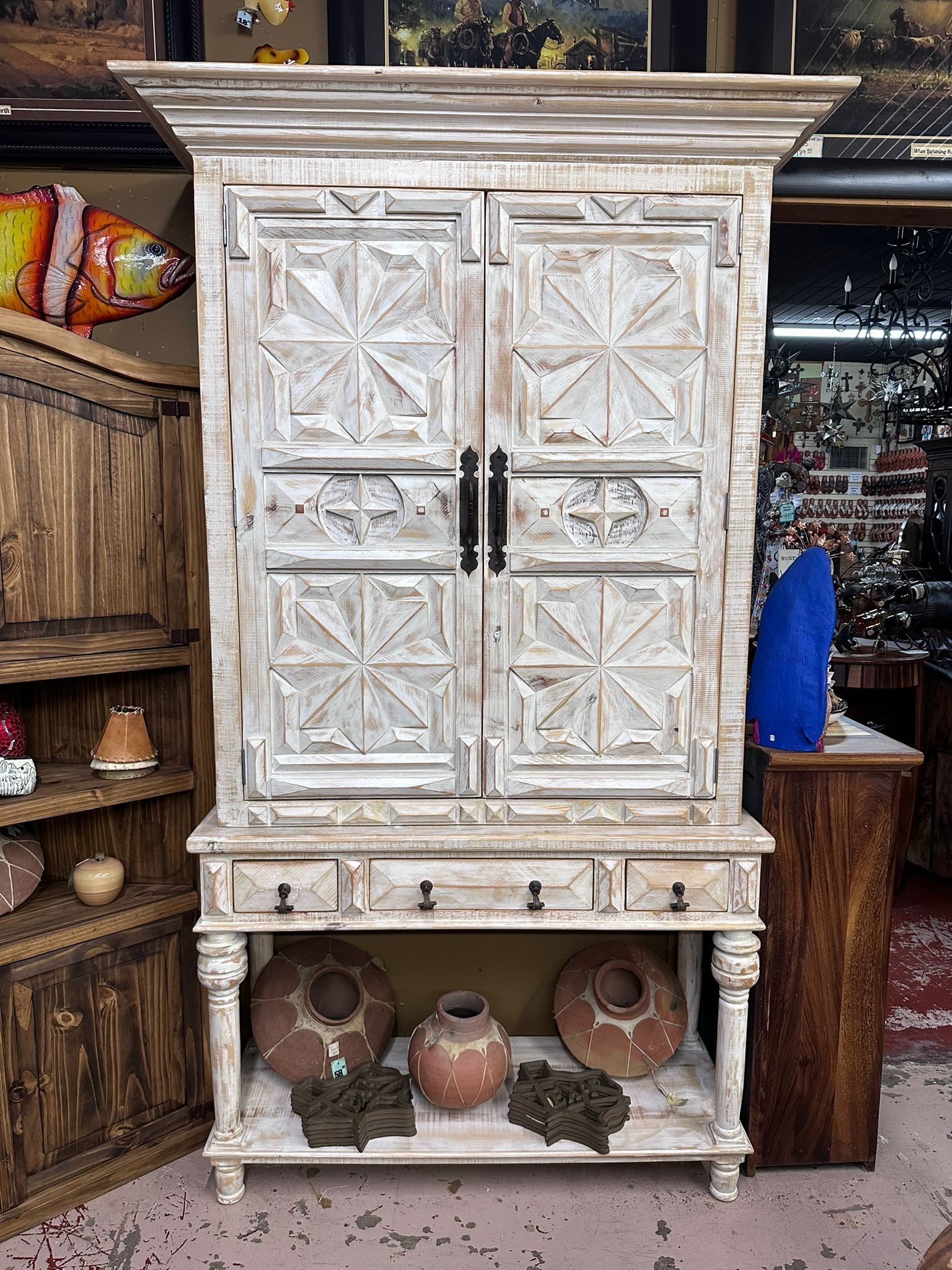 White distressed wooden cabinet with patterned doors, three drawers, and a shelf, displayed in a shop setting.