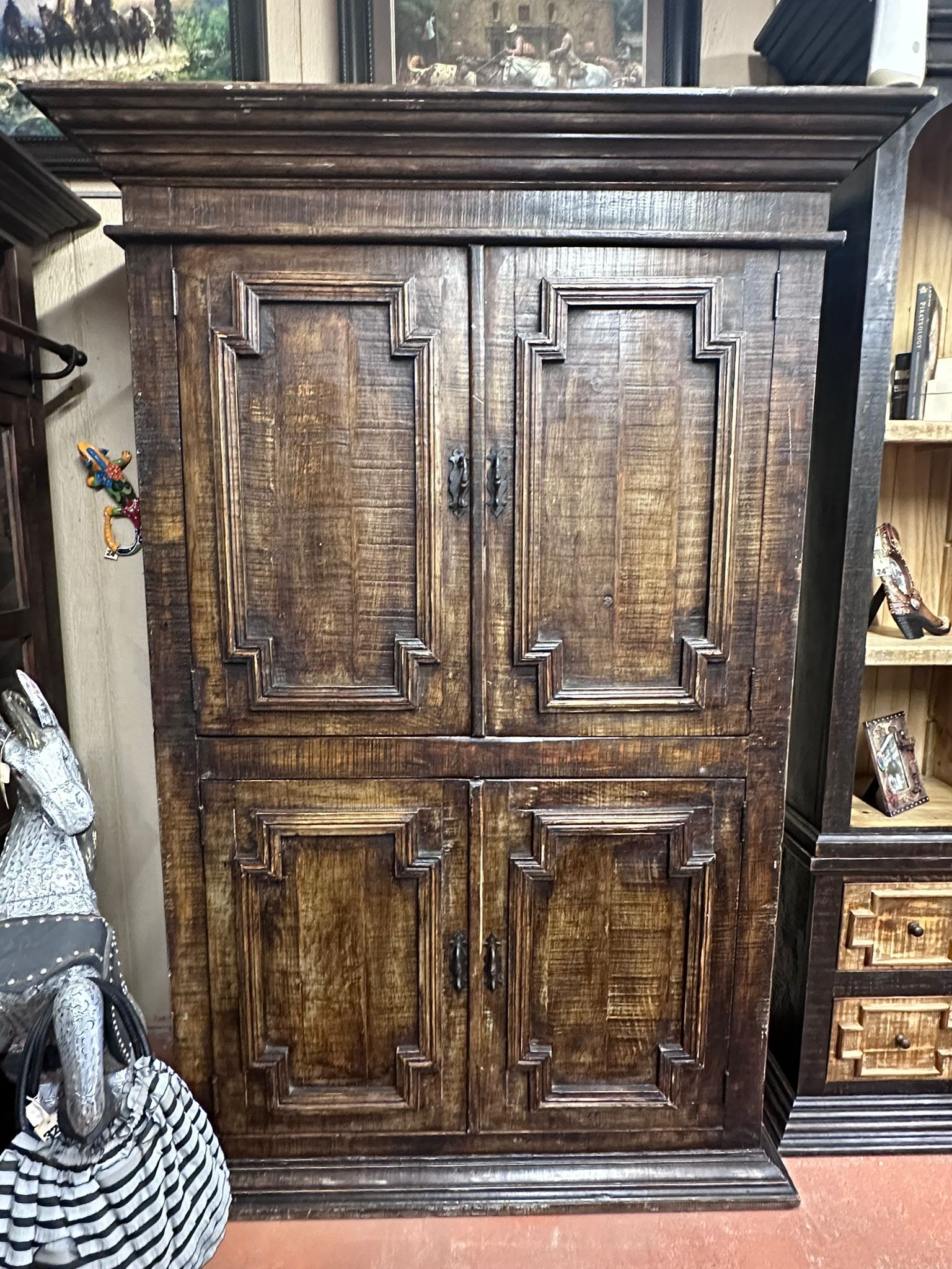 Dark wooden cabinet with four doors. It has decorative molding and a rough, aged appearance, set in a room with other wooden furniture.