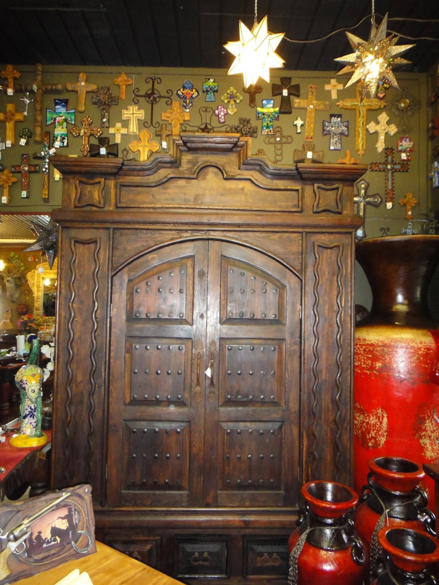 Large, dark wooden armoire with studded doors, flanked by red pottery, in a shop with crosses and star lights.