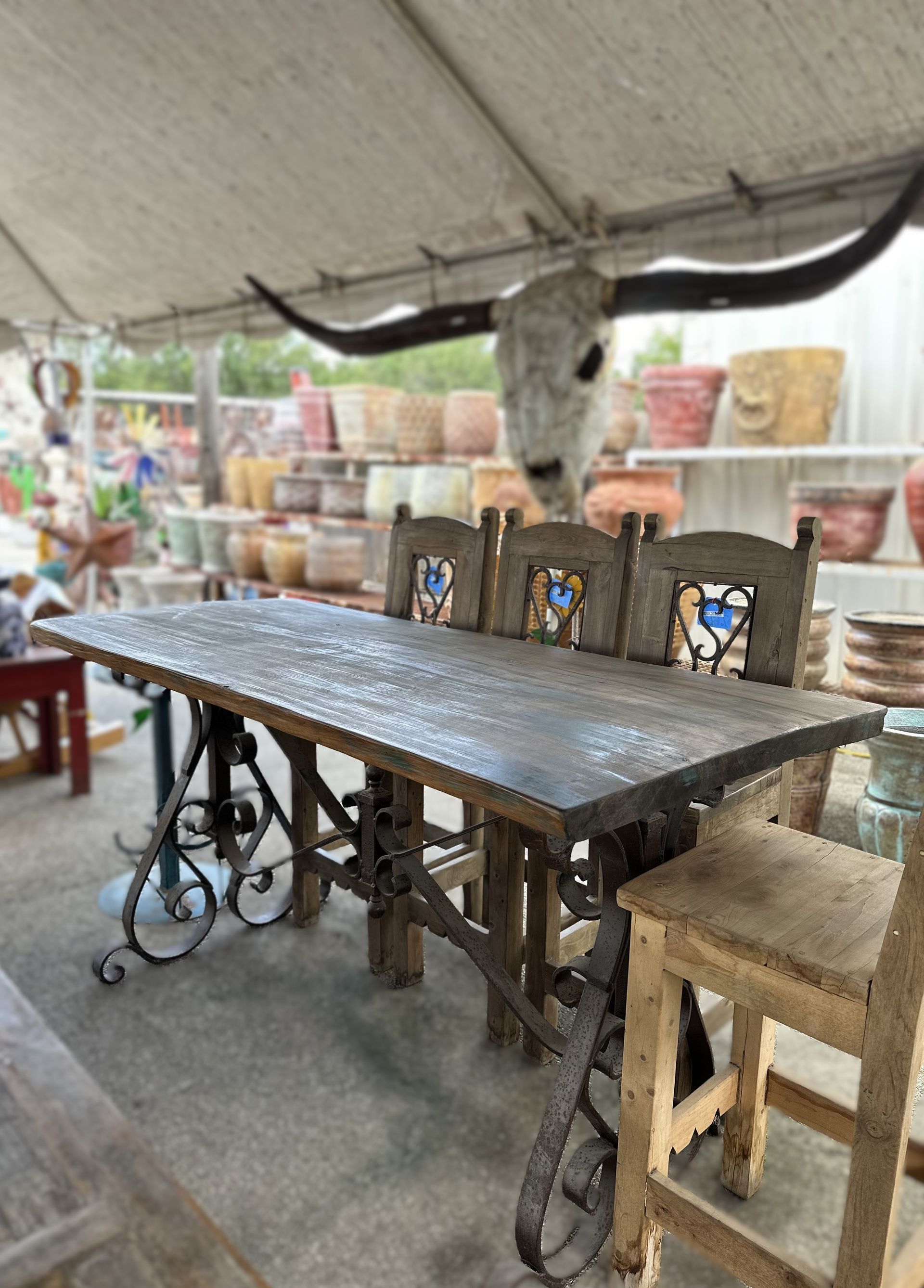 Rustic wooden table with wrought iron base and chairs, set outdoors under a canopy with a steer skull in the background.