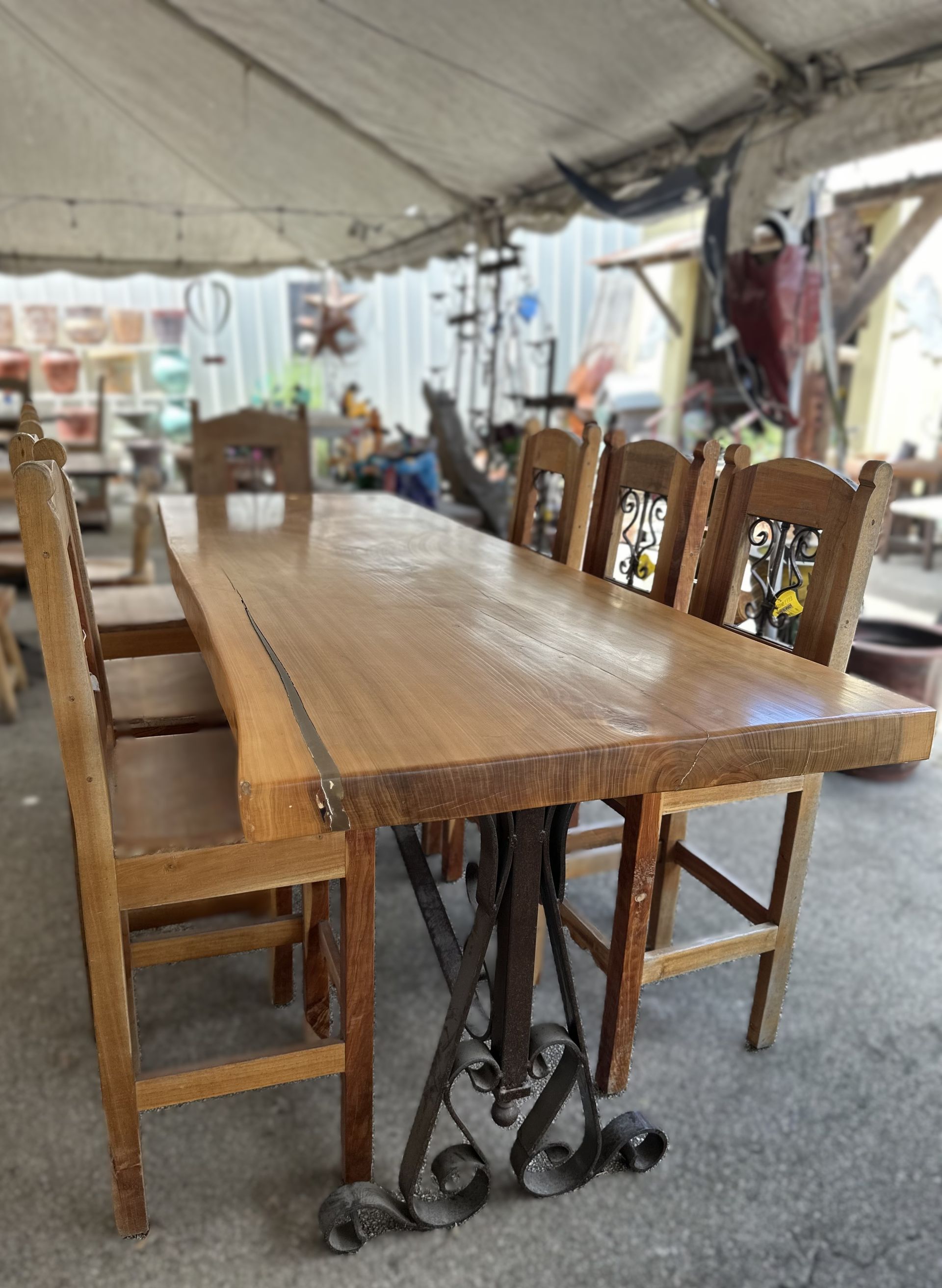 Wooden dining table with eight chairs. The table has a rustic wood top and ornate metal legs. The setting appears to be an outdoor market.