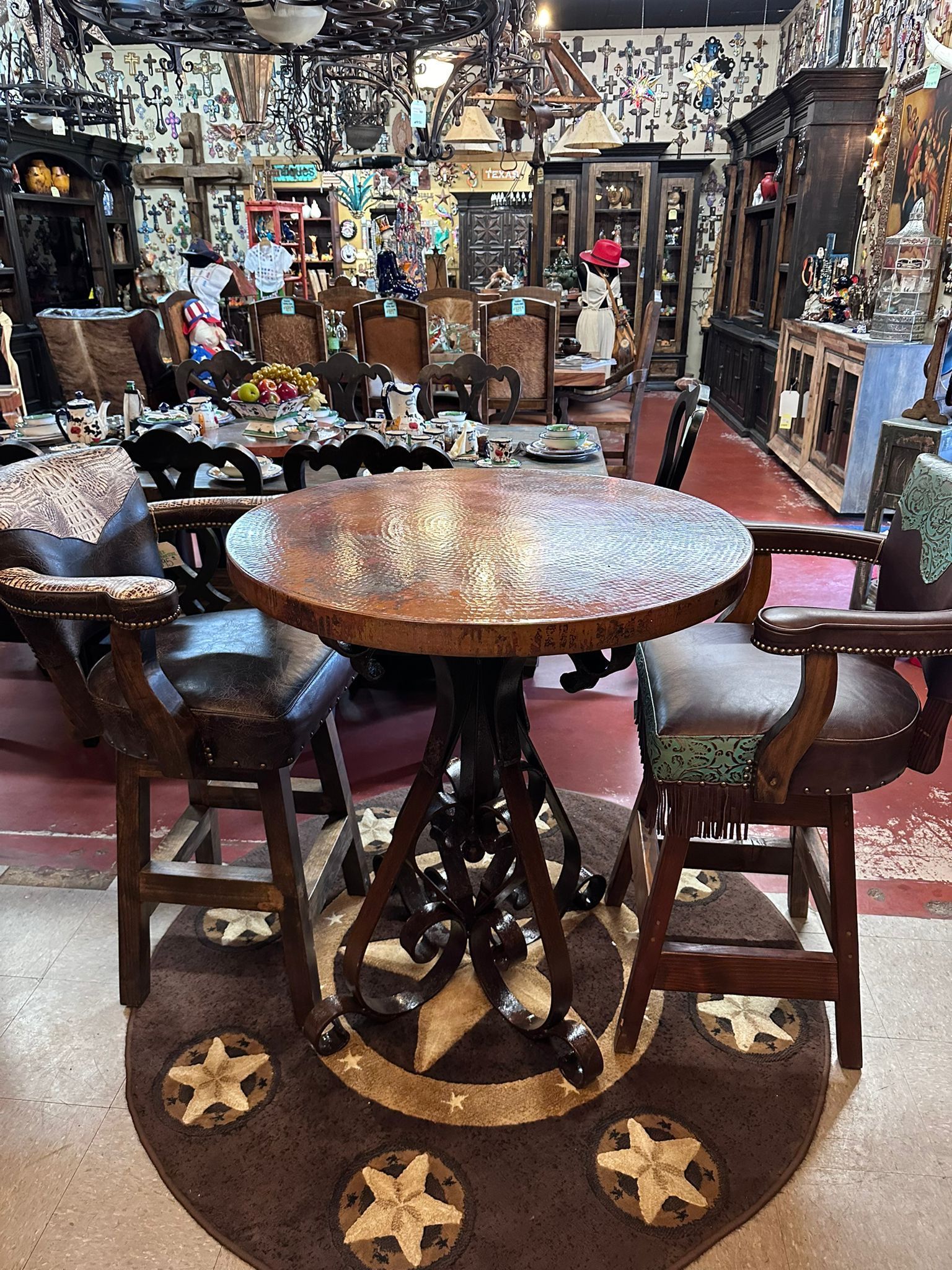 A round copper-topped table with two bar stools on a patterned rug in a cluttered room filled with furniture and decor.