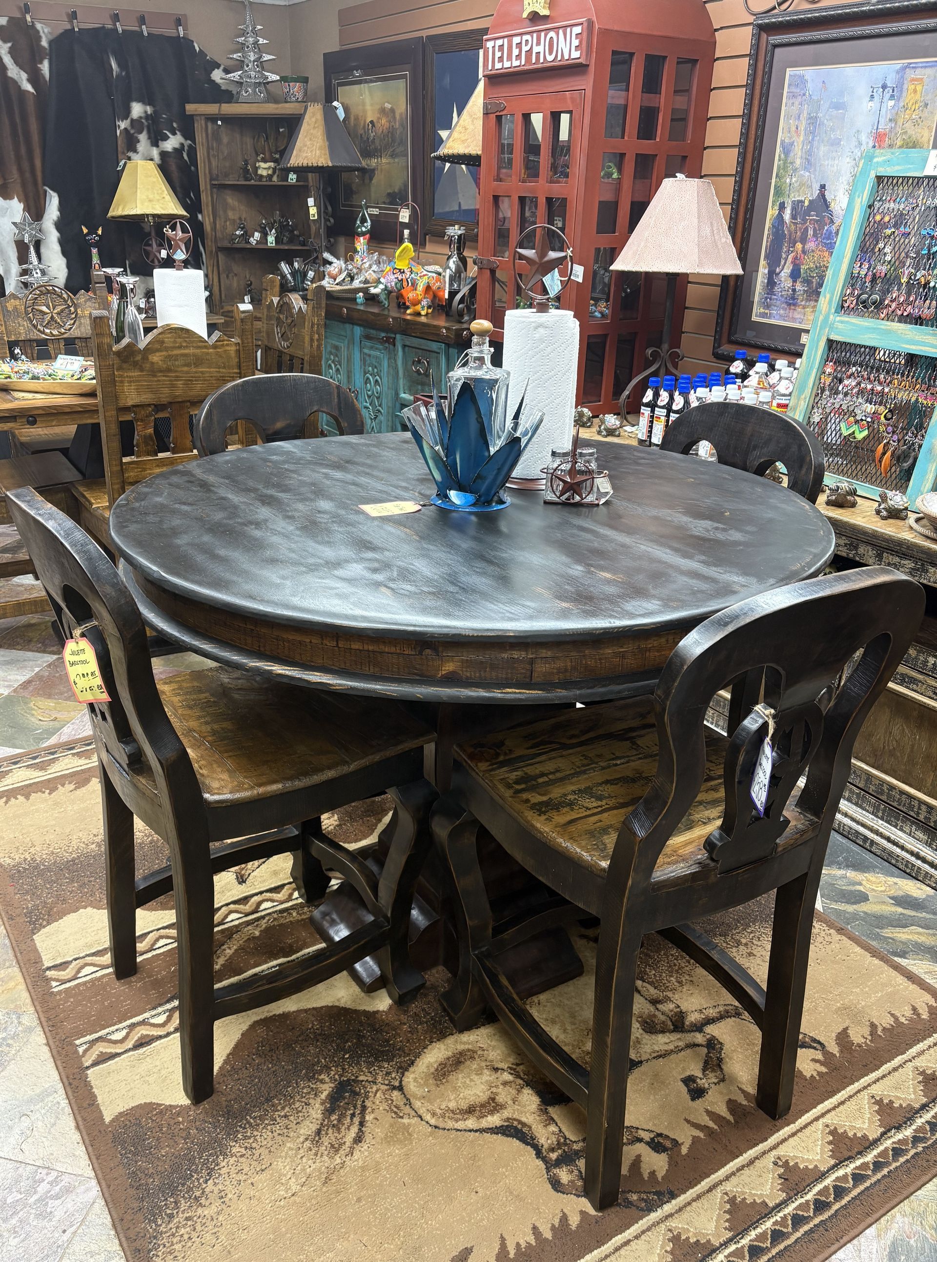 A round dark wooden dining table with four matching chairs in a rustic-style furniture store. The table is set with a decorative bottle and items.