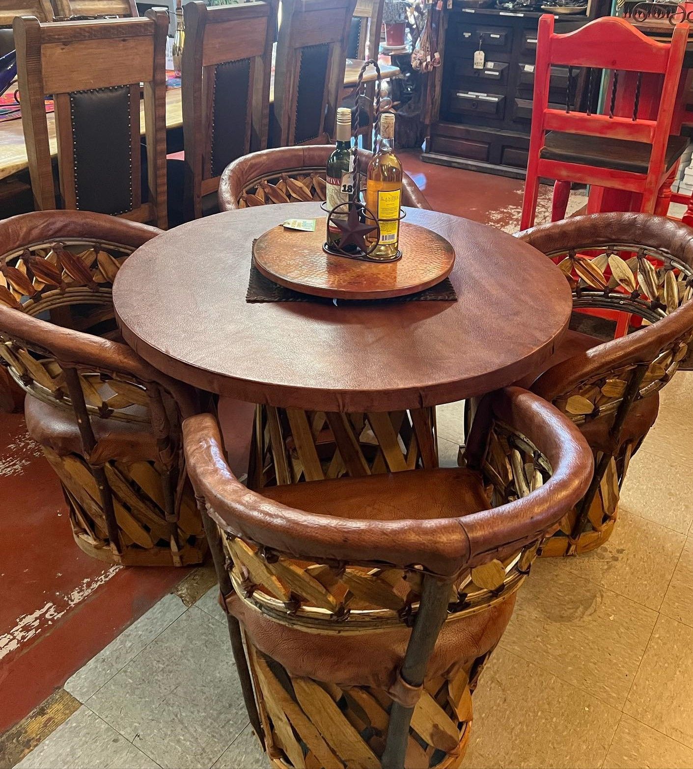 A wooden table and four matching leather woven chairs. Bottles sit on the table's center, in a shop setting.