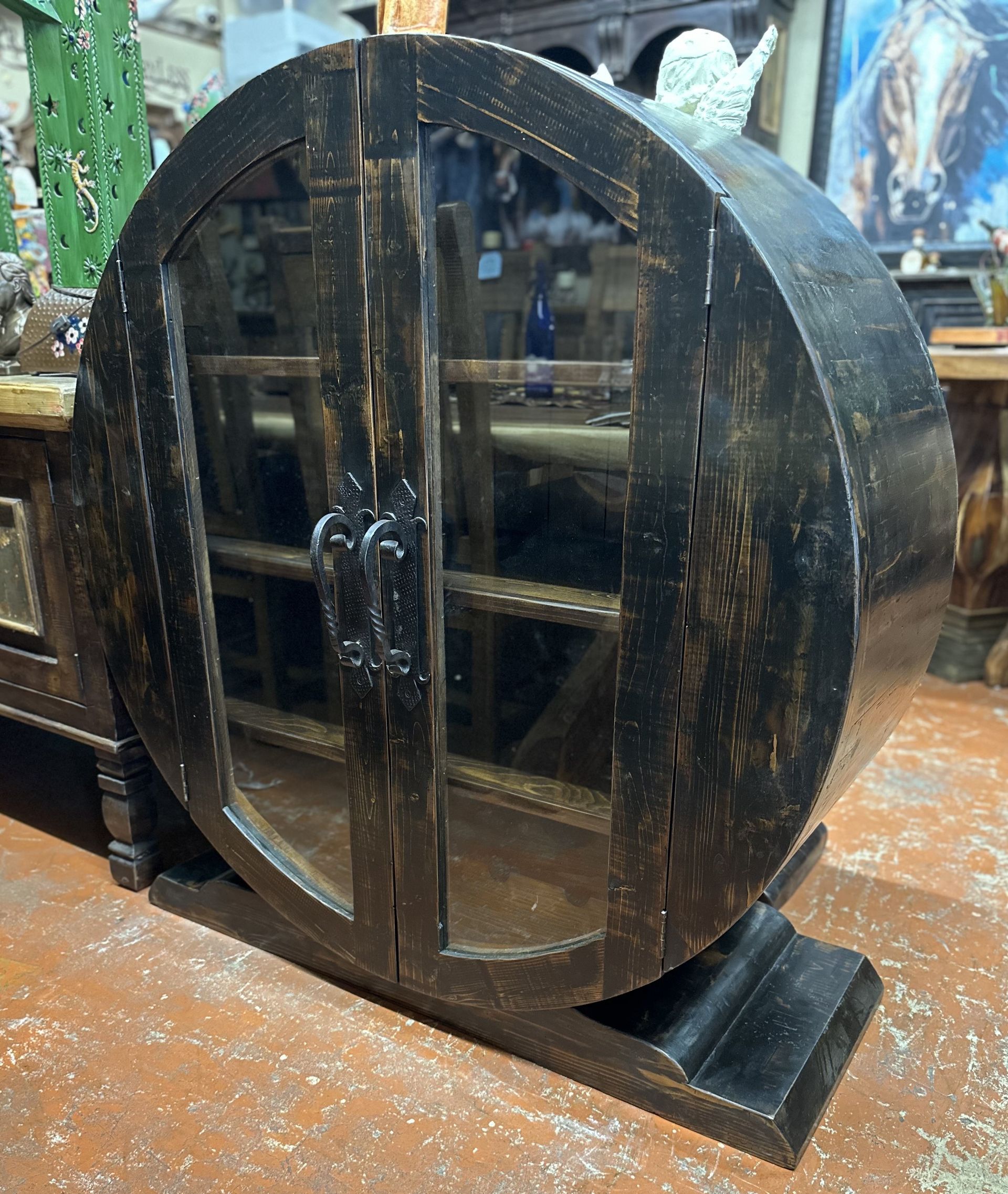 A rustic, barrel-shaped cabinet with glass doors, painted black. It sits on a matching base, and the shelves are visible inside.