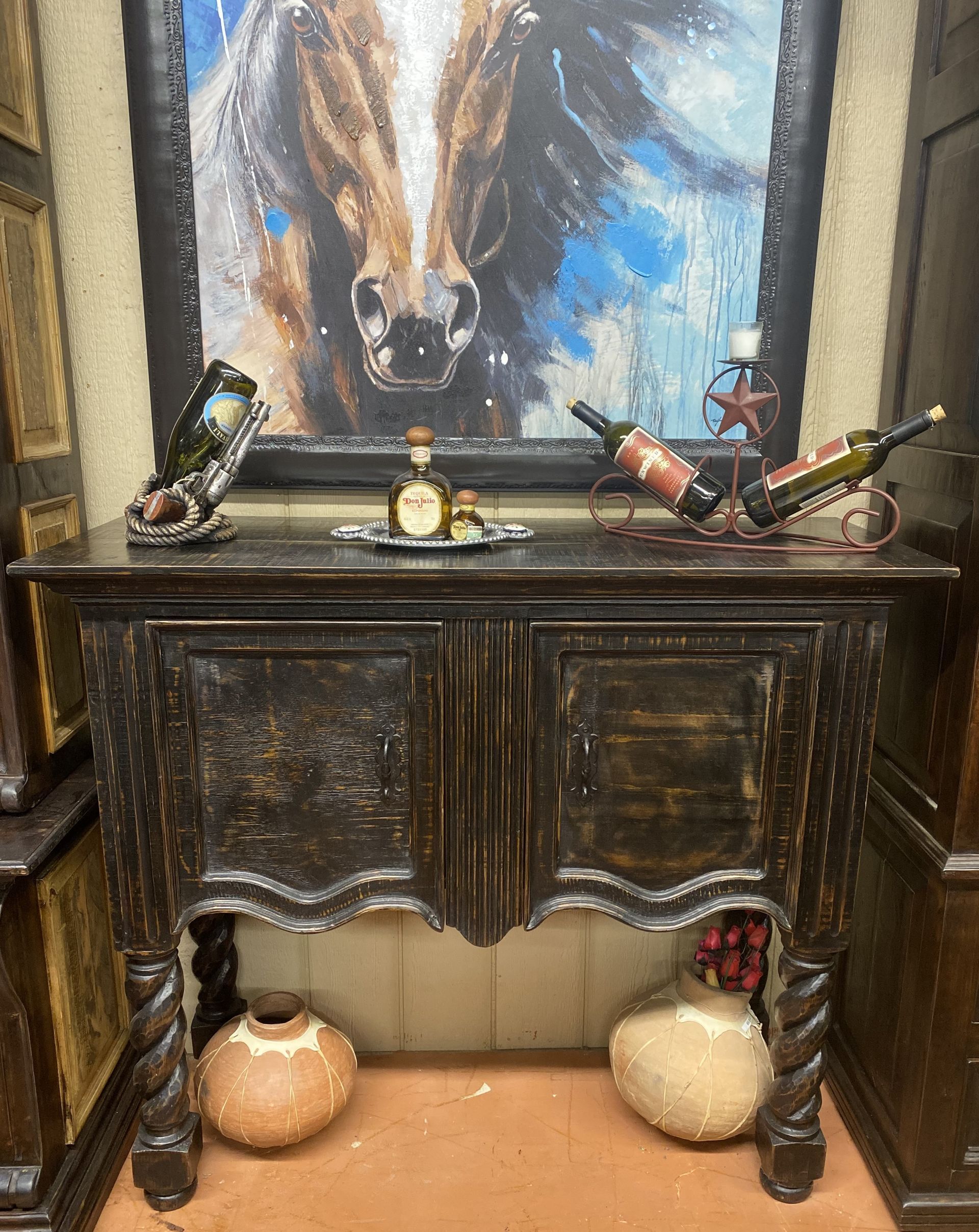 A dark wood sideboard with wine bottles, artwork of a horse, and decorative pottery.
