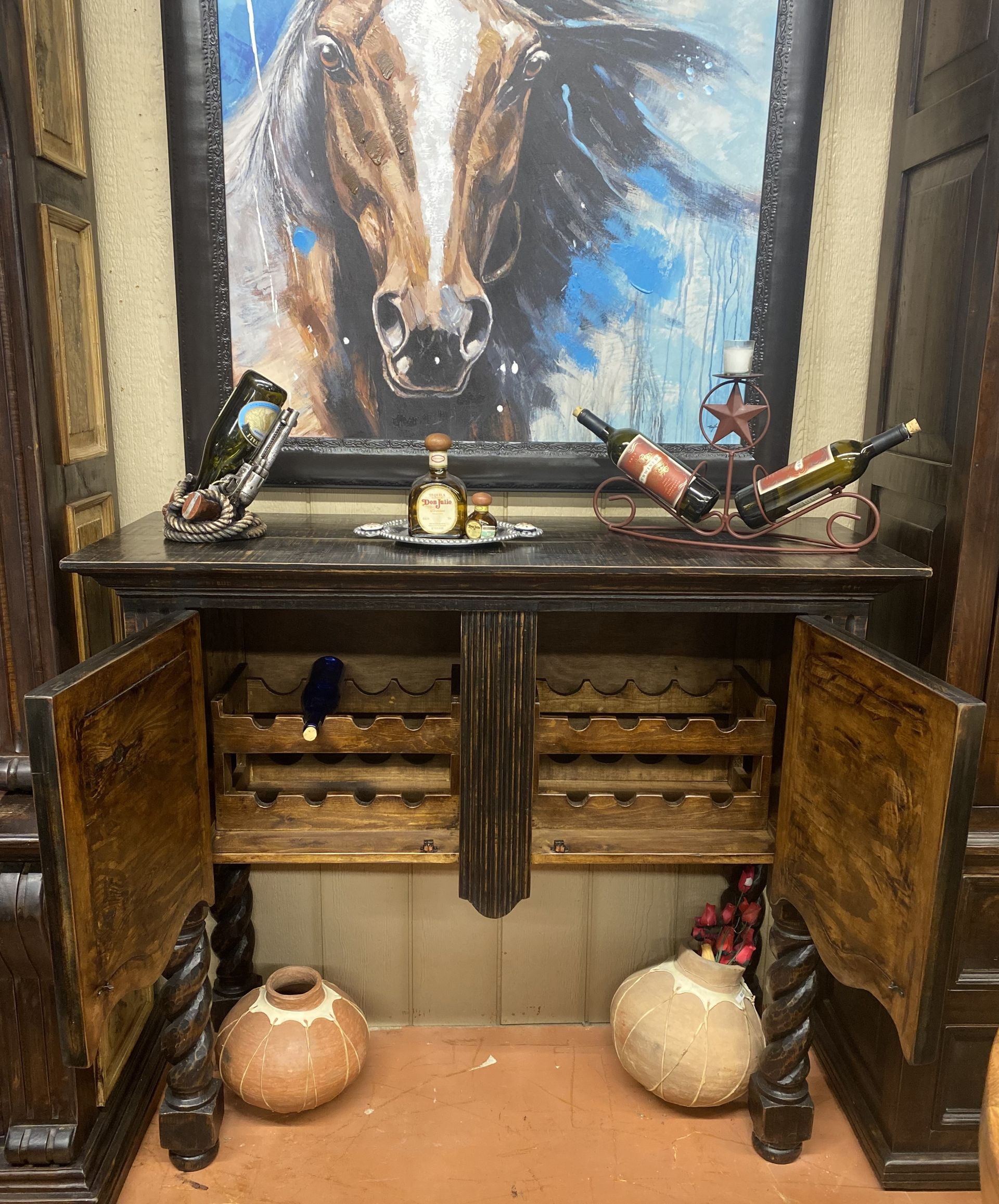 A wooden wine cabinet with open doors, holding bottles and decorative pottery. A painting of a horse hangs above the cabinet.