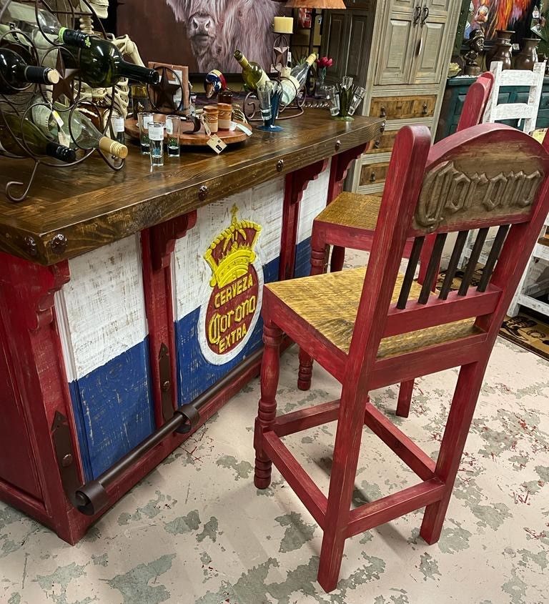 Red, rustic bar with Corona logo, two matching bar stools with woven seats, and various bottles on the counter.