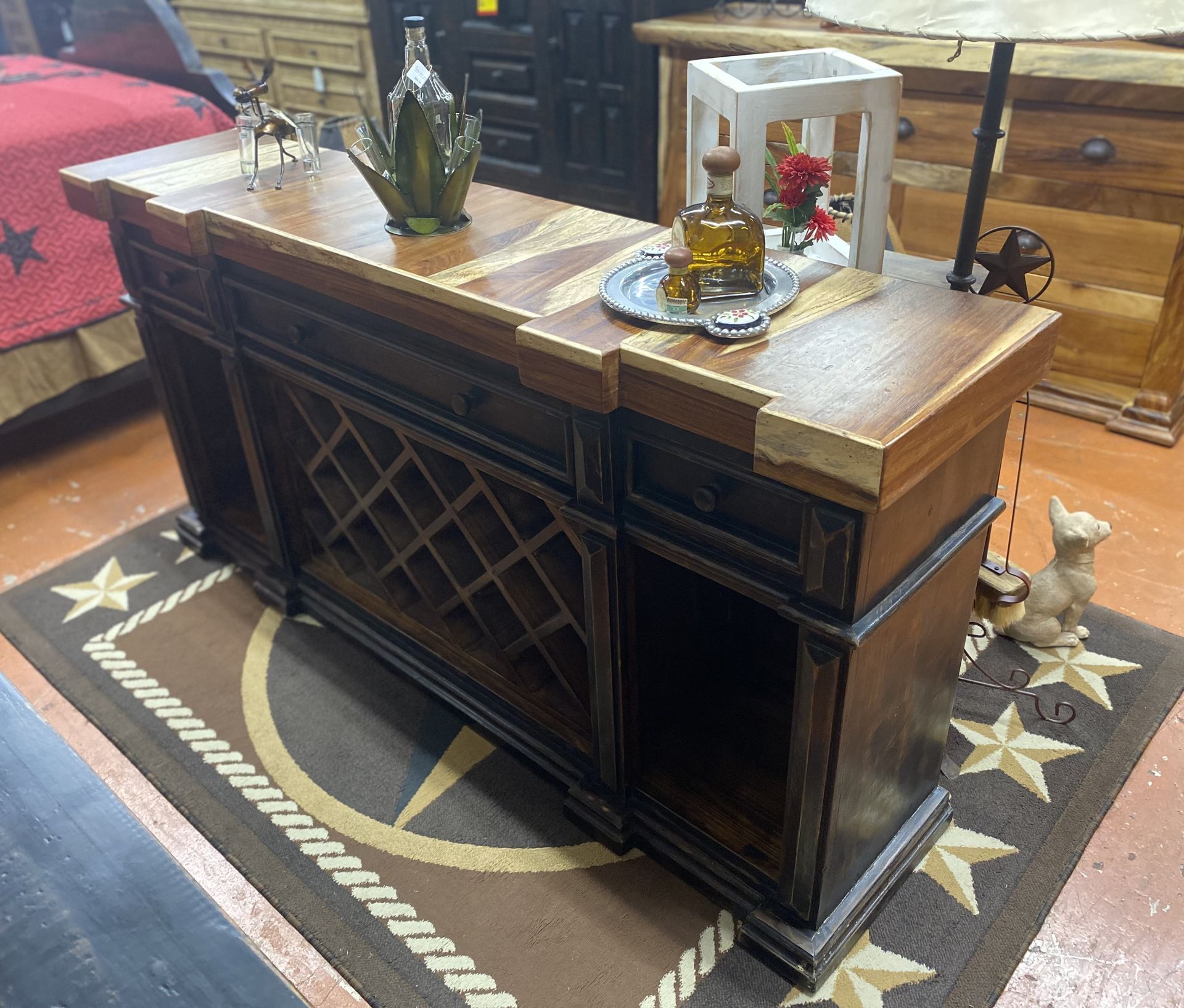 A rustic wooden bar with a wine rack, liquor bottles, and a metal star accent. Set on a star-patterned rug.