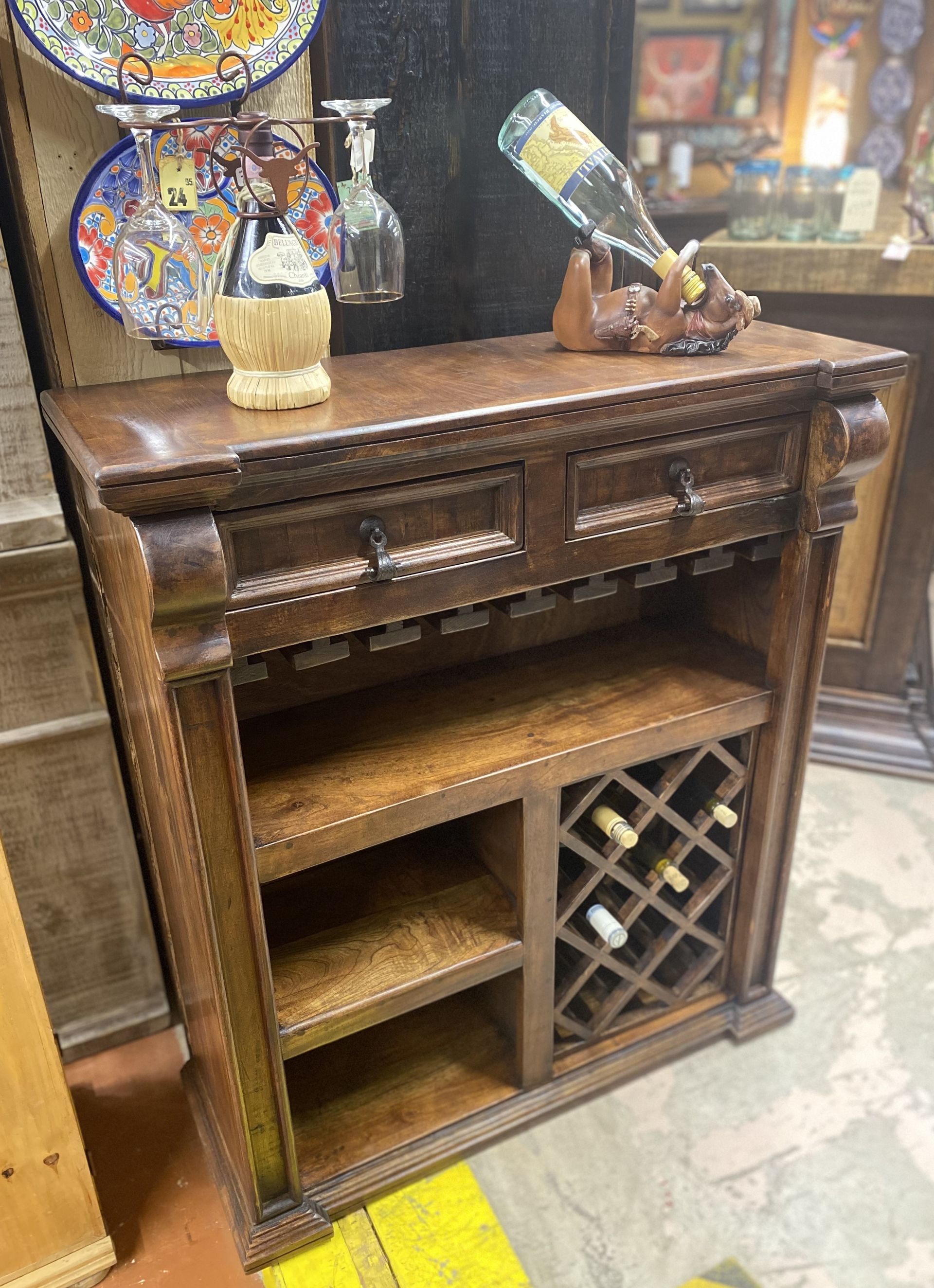 Wooden bar cabinet with a wine rack, drawers, and shelf space. There is a bottle of wine and glasses displayed on the top.