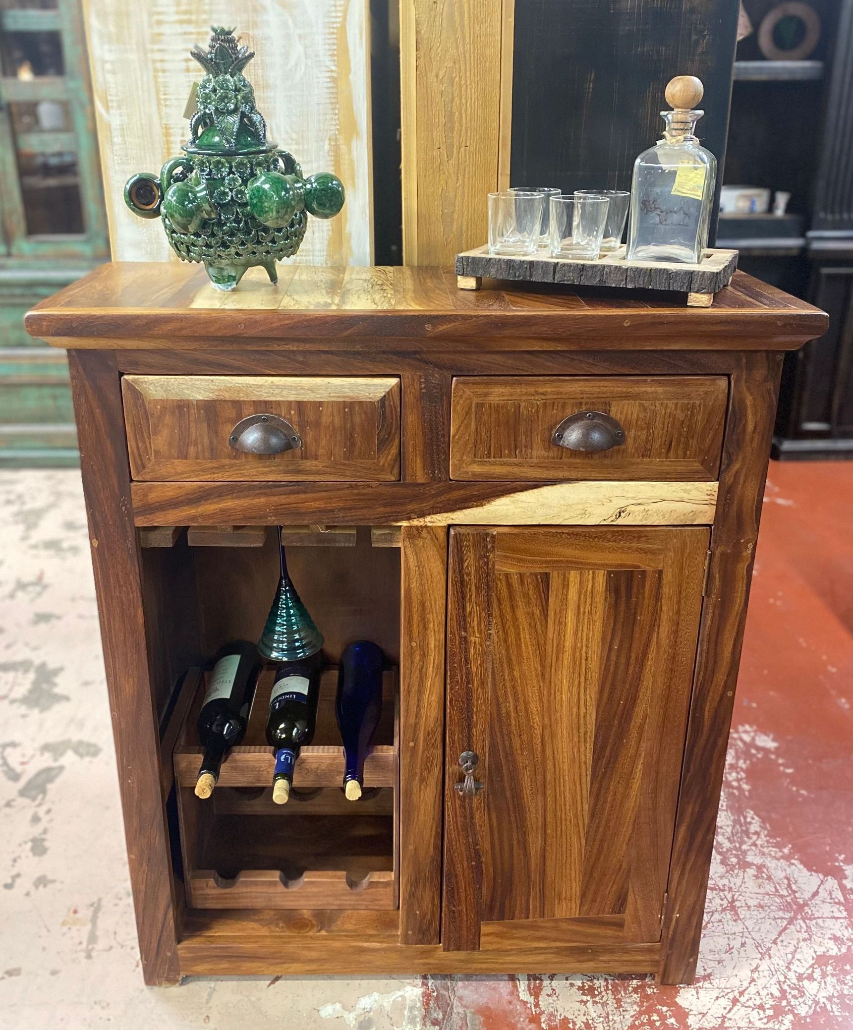 Wooden mini bar with drawers, a cabinet door, and wine storage, topped with a decorative object and a tray of shot glasses and a bottle.