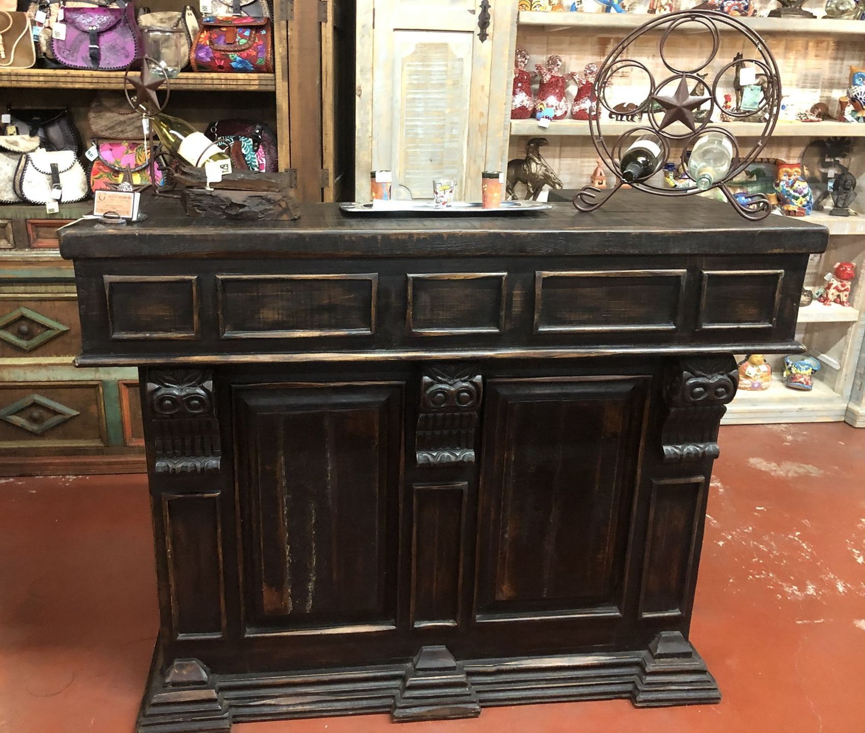 Dark wooden store counter with intricate carvings. A metal wine rack and merchandise are on the counter and shelves are behind it.