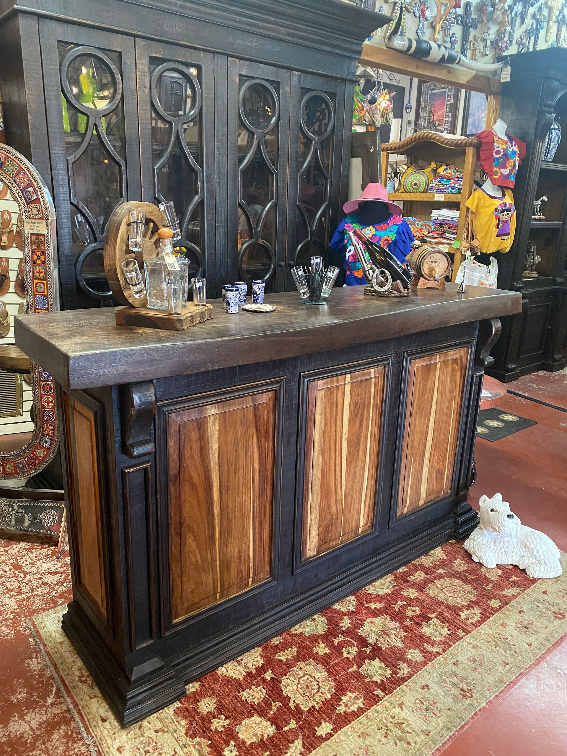 A detailed wooden bar with a dark finish, showcasing ornate carvings and a textured countertop. Merchandise and a rug are around it.
