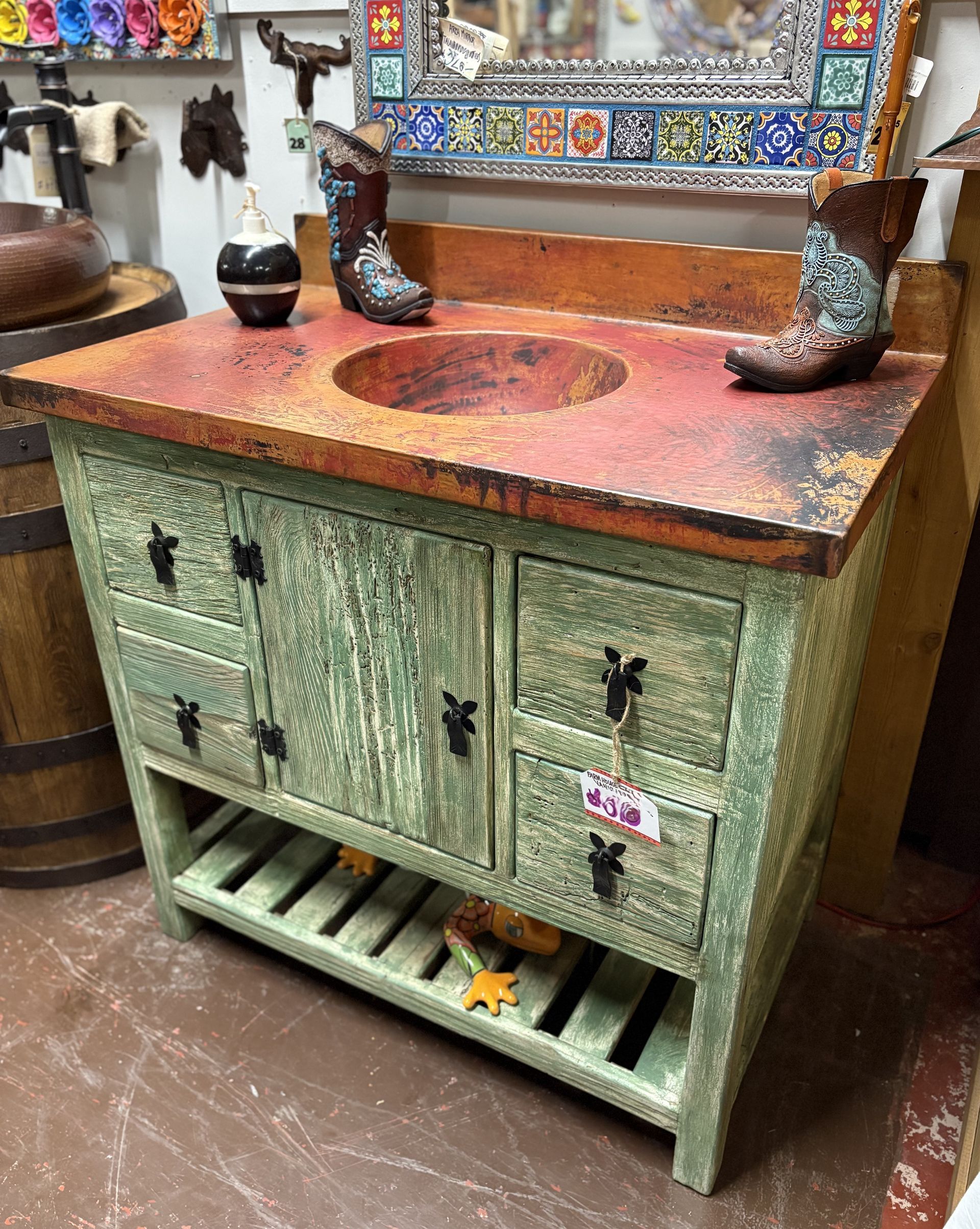 Rustic wooden bathroom vanity with a copper sink and countertop, painted green with a weathered finish. Two cowboy boots sit on top.