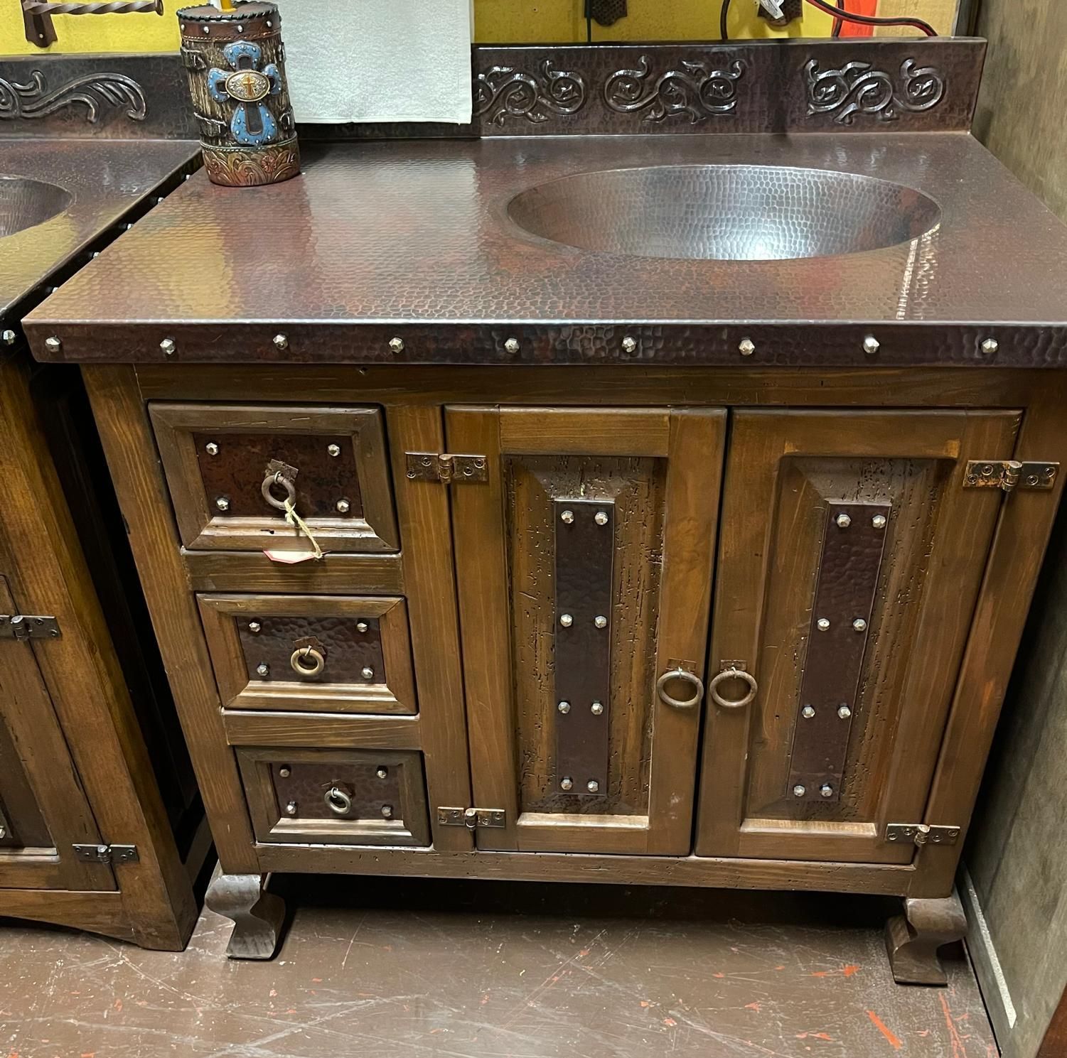 A rustic wooden bathroom vanity with a copper countertop and sink. The cabinet has drawers, doors, and decorative metal studs.