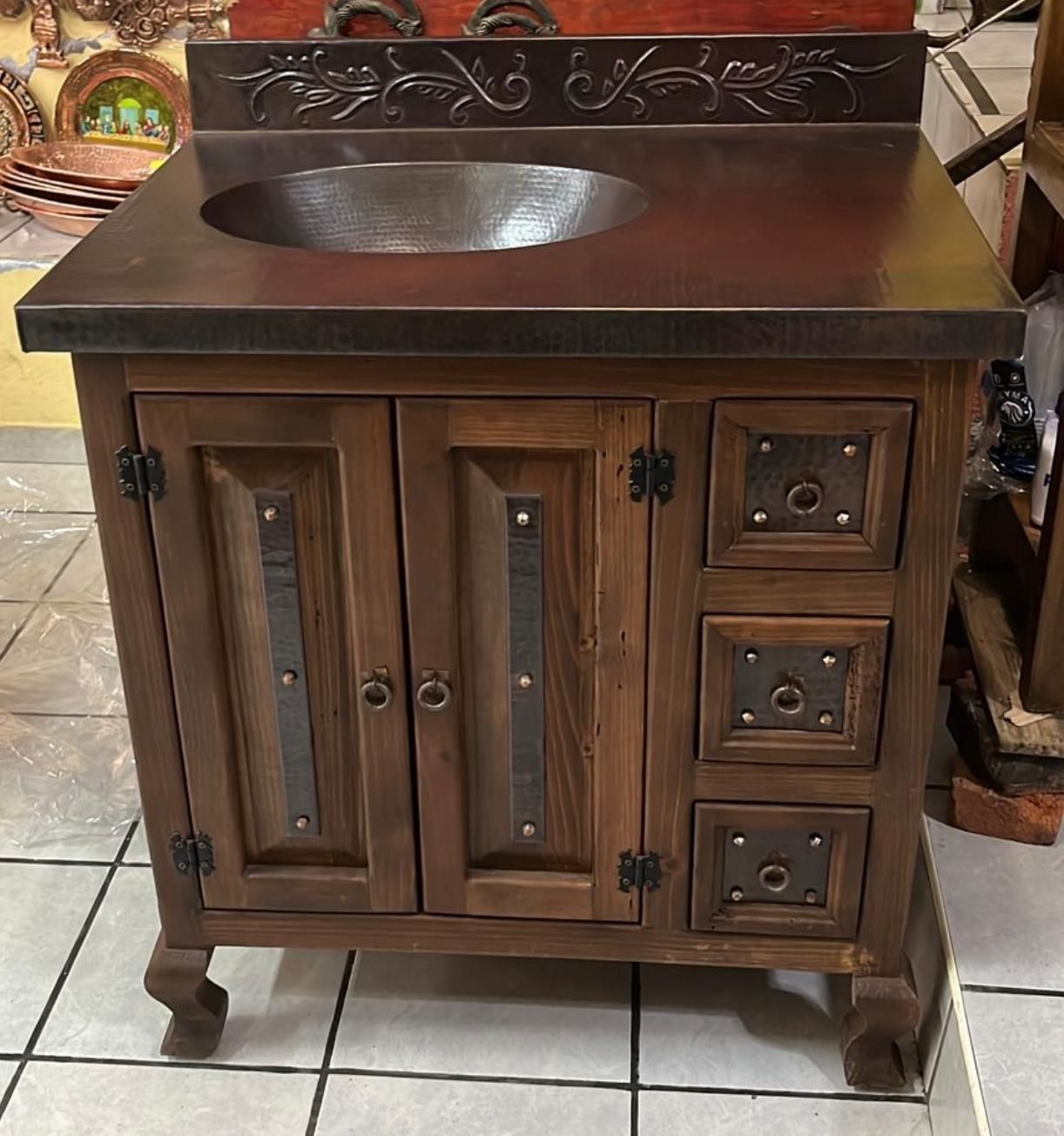 Rustic wooden bathroom vanity with copper sink and decorative metal accents. It has two doors and three drawers.