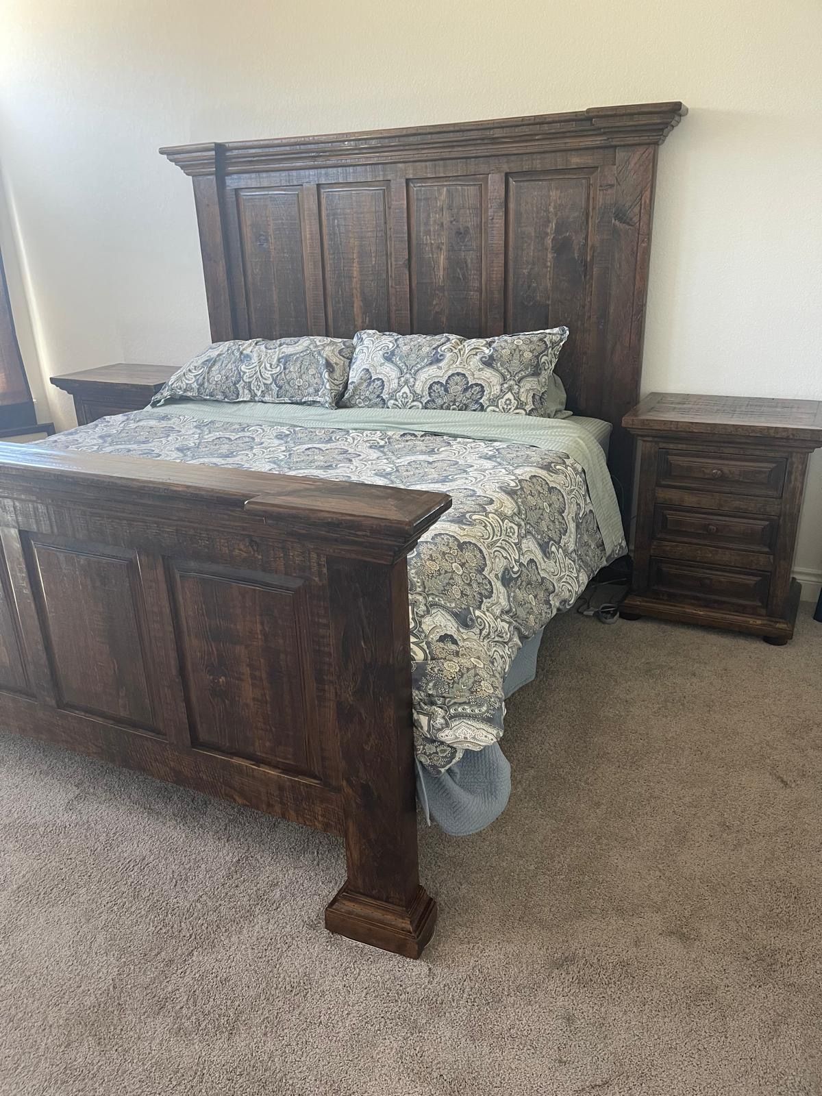 A rustic wooden bed with matching nightstands, covered with a floral patterned comforter, sits in a room with beige carpet.