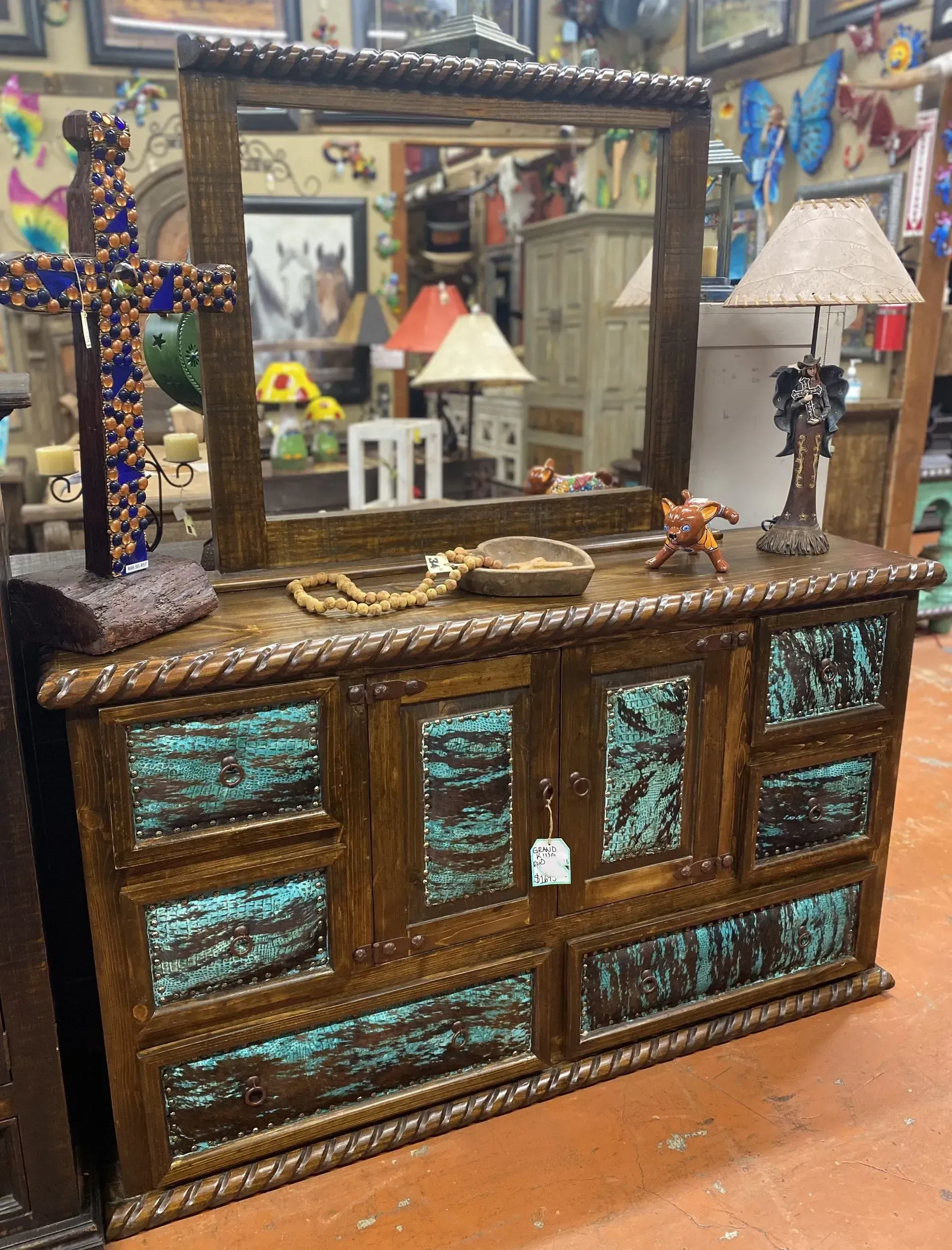 Wooden dresser with a turquoise-colored design, a rope-like trim, and a mirror. There are decorative items on top, like a cross and a lamp.