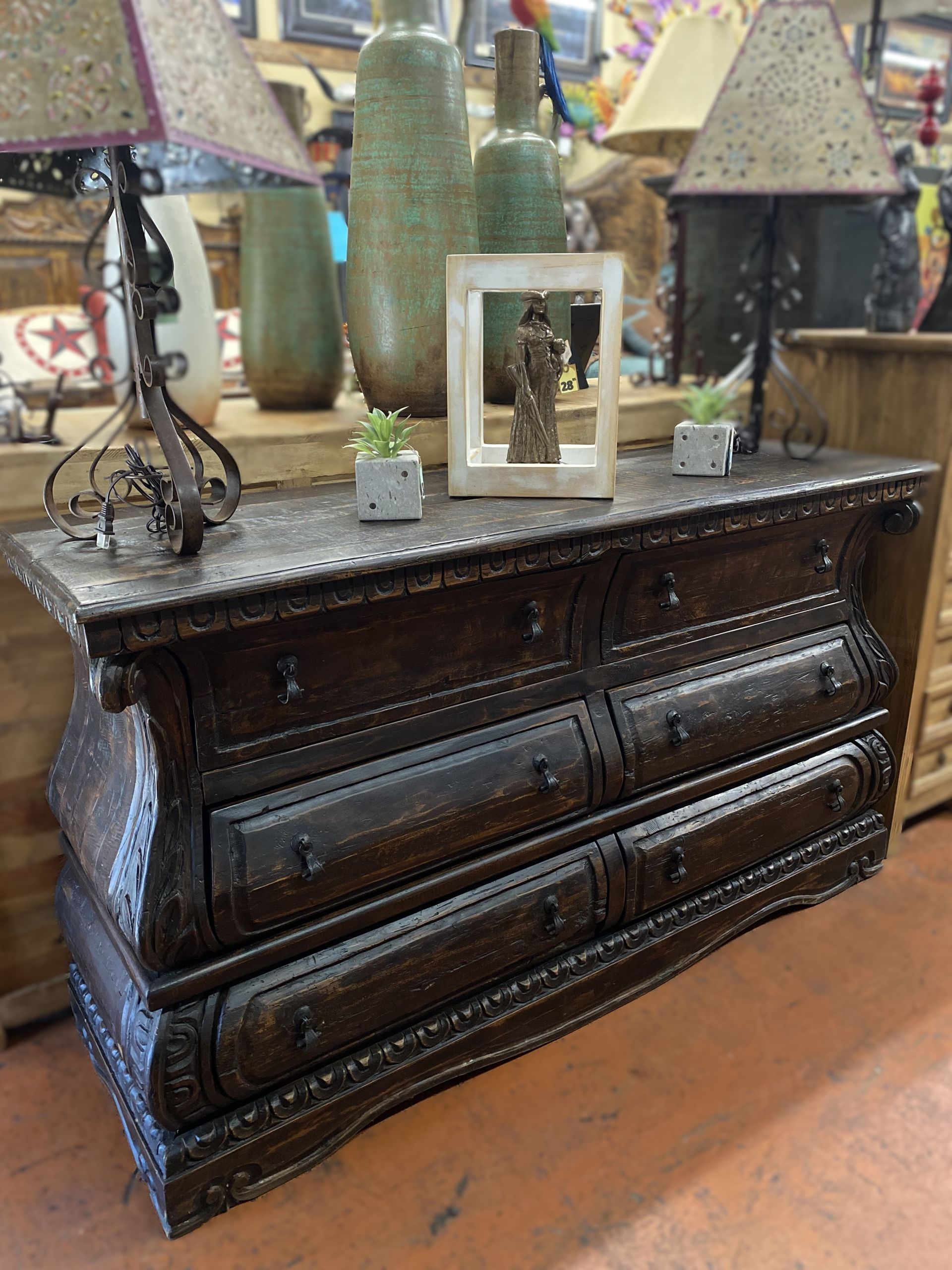 Dark wooden dresser with six drawers, topped with vases, lamps, and small plants. Displayed in a shop setting.