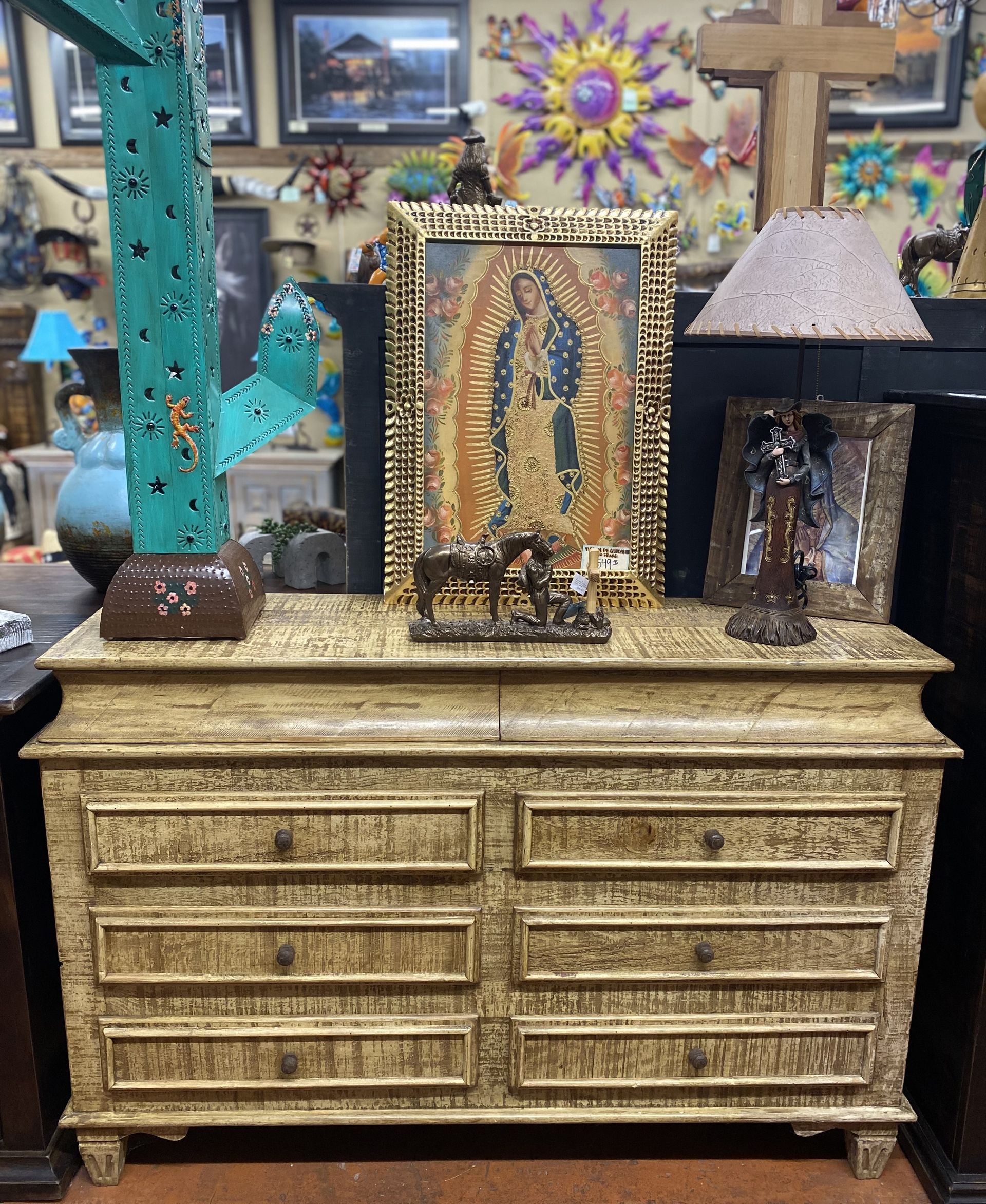 A beige dresser displays religious artwork, including a gold-framed painting and statues. The setting is a cluttered shop with other decor.
