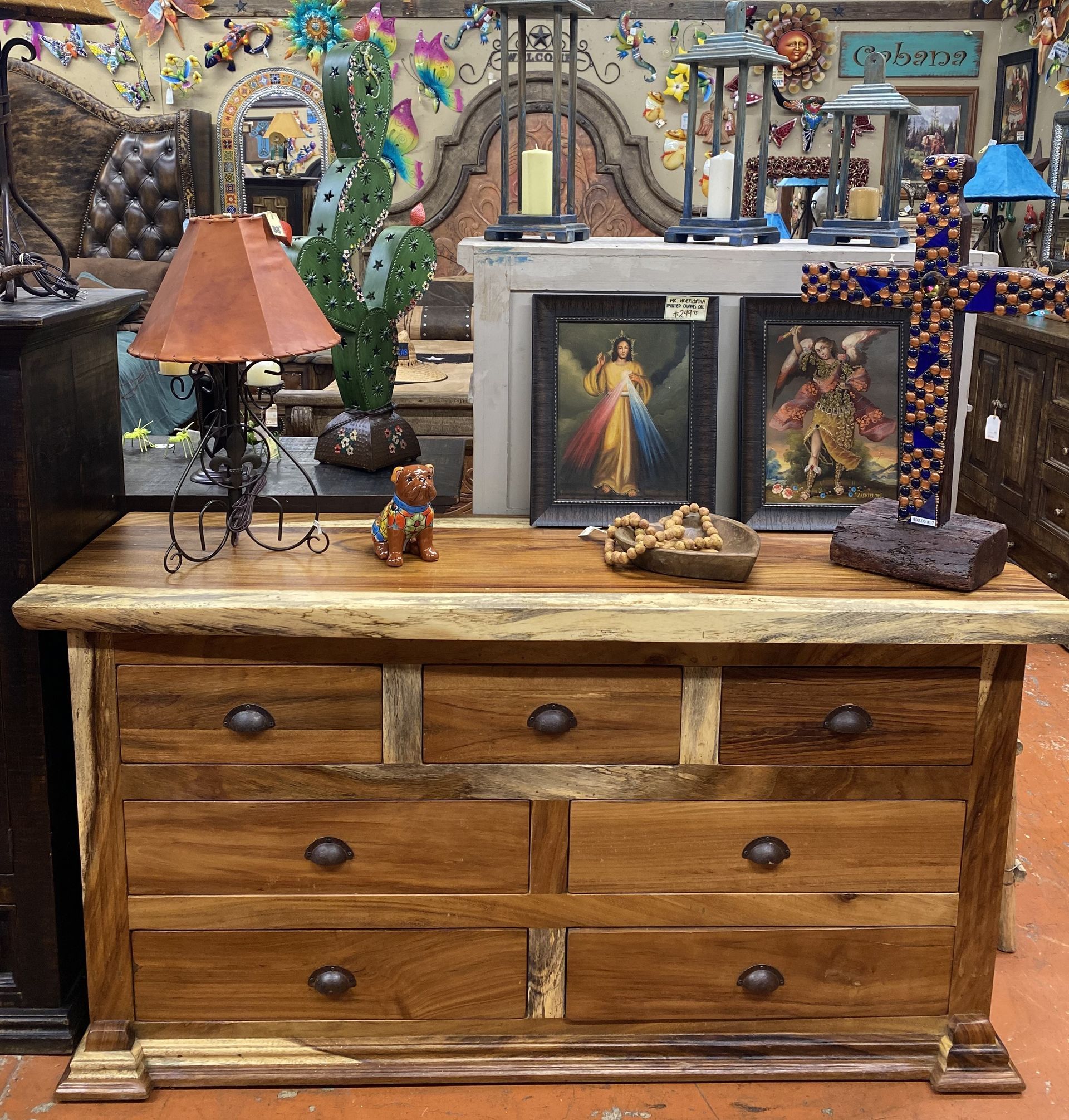 Wooden dresser with seven drawers. Interior with religious art, a lamp, and cactus.