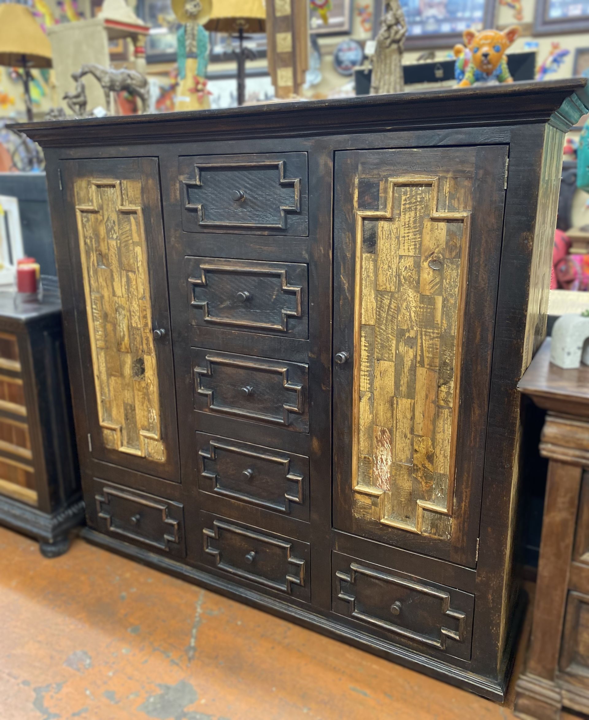 Dark wooden cabinet with gold-colored accents. It has doors, drawers, and intricate carvings. The setting appears to be a furniture store.