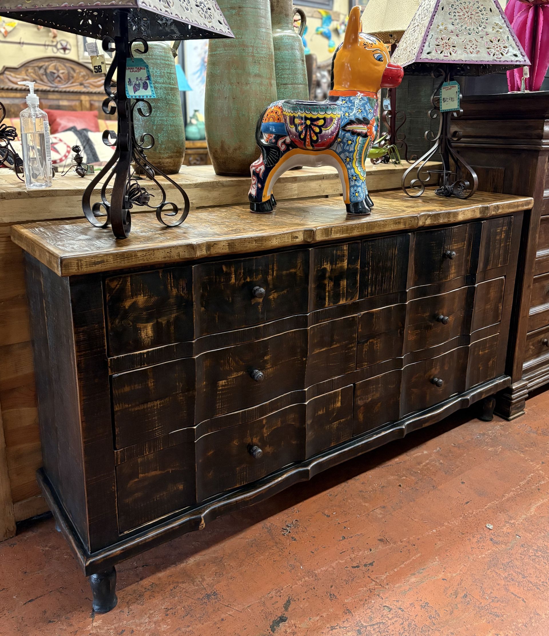 A dark wood dresser with multiple drawers, adorned with metal accents and topped with decorative items like lamps and a colorful donkey figurine, set in a shop.