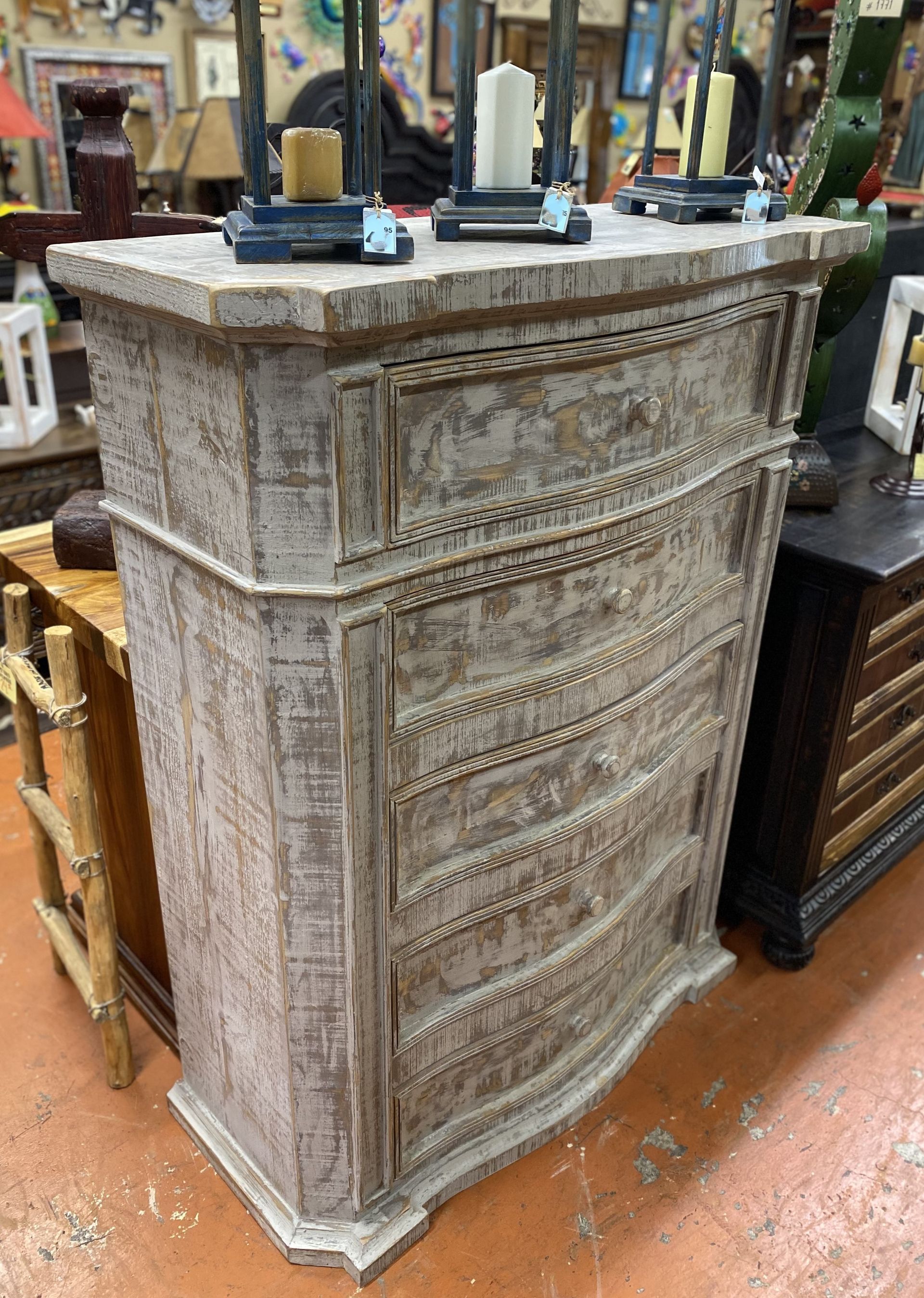 A distressed, light-colored chest of drawers with curved drawers. The top surface holds candles. It's in a store setting.