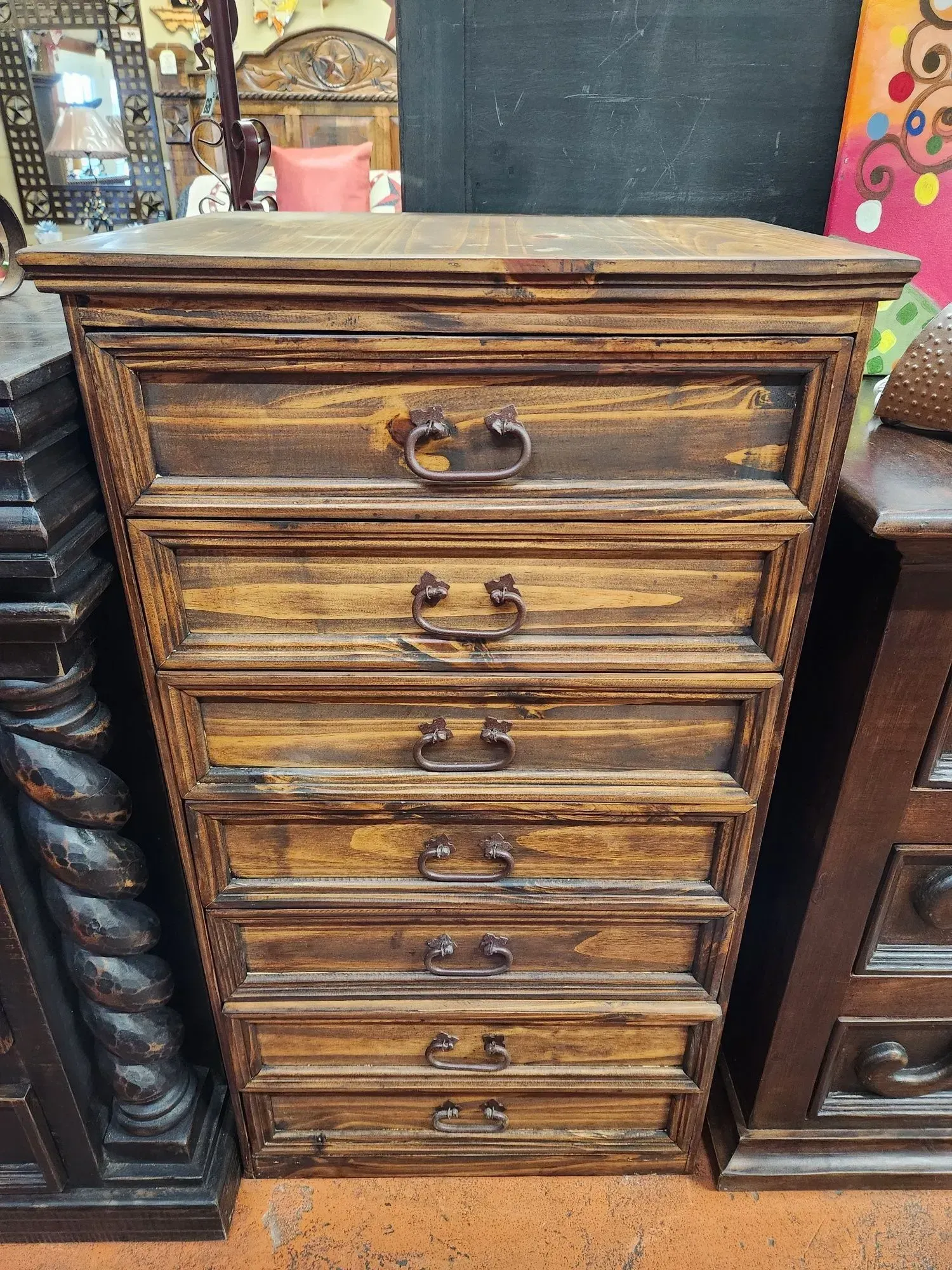Wooden chest of drawers with six drawers and ornate pulls, stained dark brown. The drawers are set in a room with other furniture.