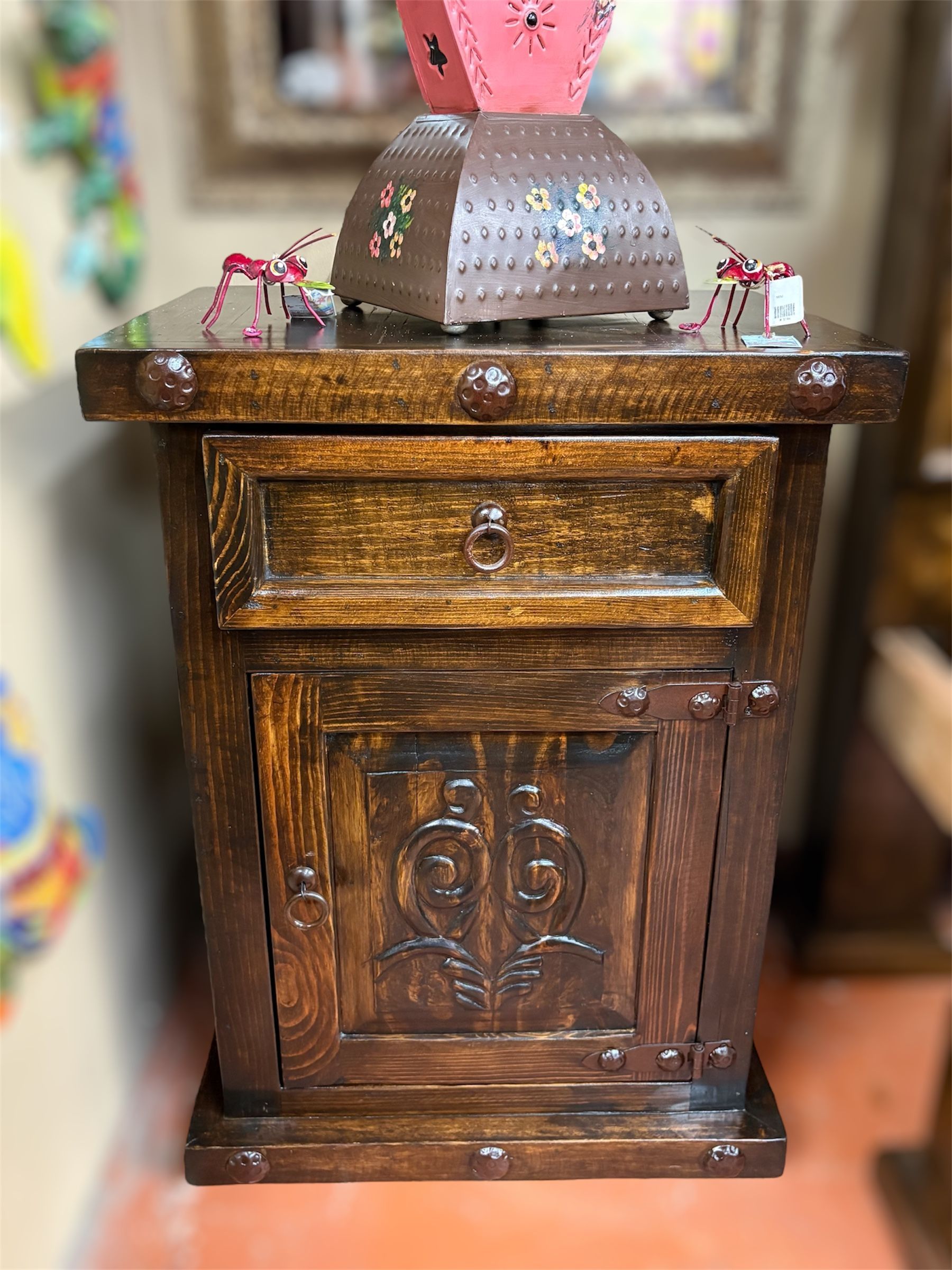 Wooden nightstand with a drawer and cabinet. It is dark brown with decorative carvings and metal studs. A decorative object sits on top.