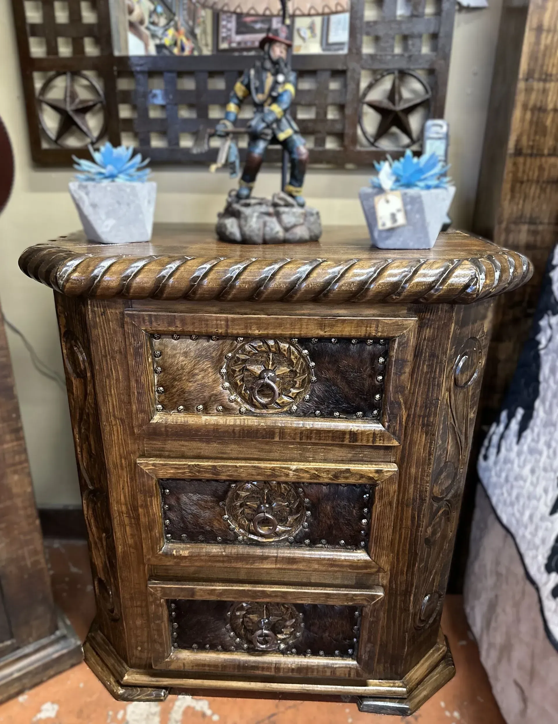 Wooden nightstand with three drawers, rope-like trim, and decorative metal accents.  Two blue succulents and a figurine top it.
