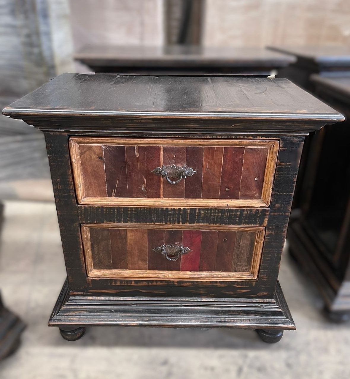 Rustic wooden nightstand with two drawers and a distressed black finish. The drawer fronts are composed of vertical wood panels in various colors.
