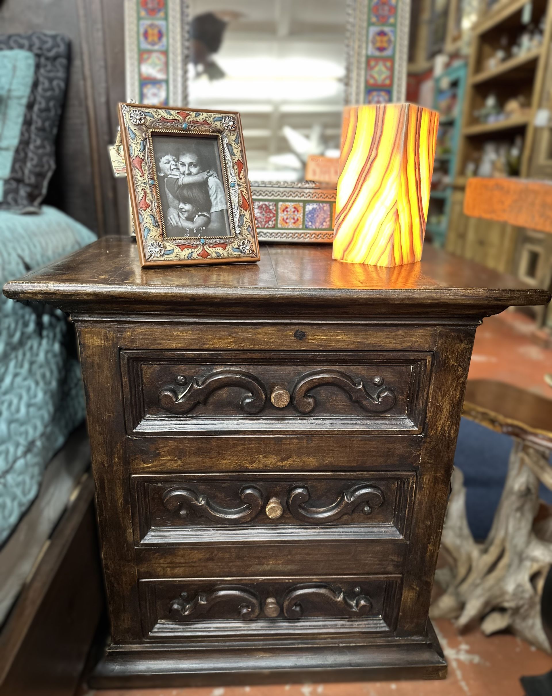 Dark wooden nightstand with three drawers, holding a photo frame and a glowing orange lamp.  The room has a rustic, warm vibe.