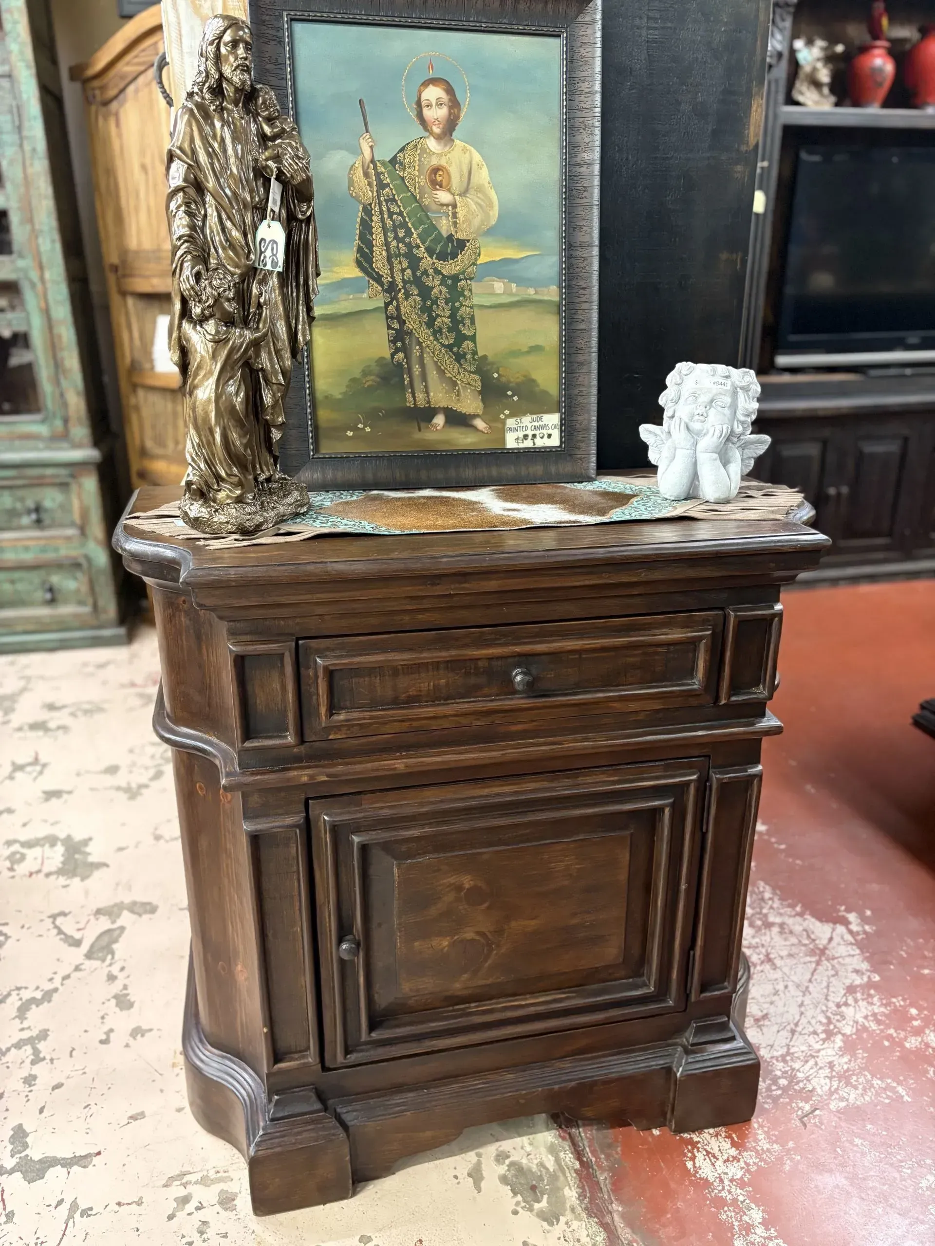Dark wooden nightstand with a drawer and cabinet, topped with religious statues and a framed picture.