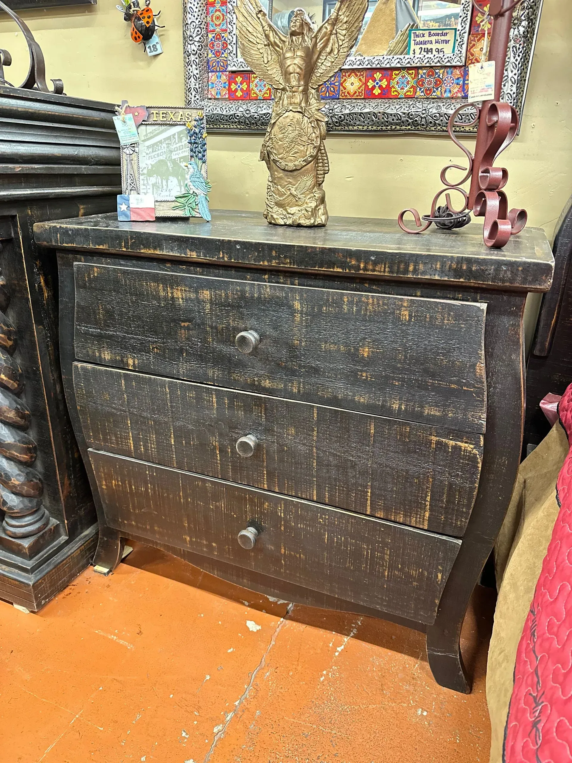 A distressed dark wood dresser with three drawers. There is a decorative statue and other items on top of the dresser.