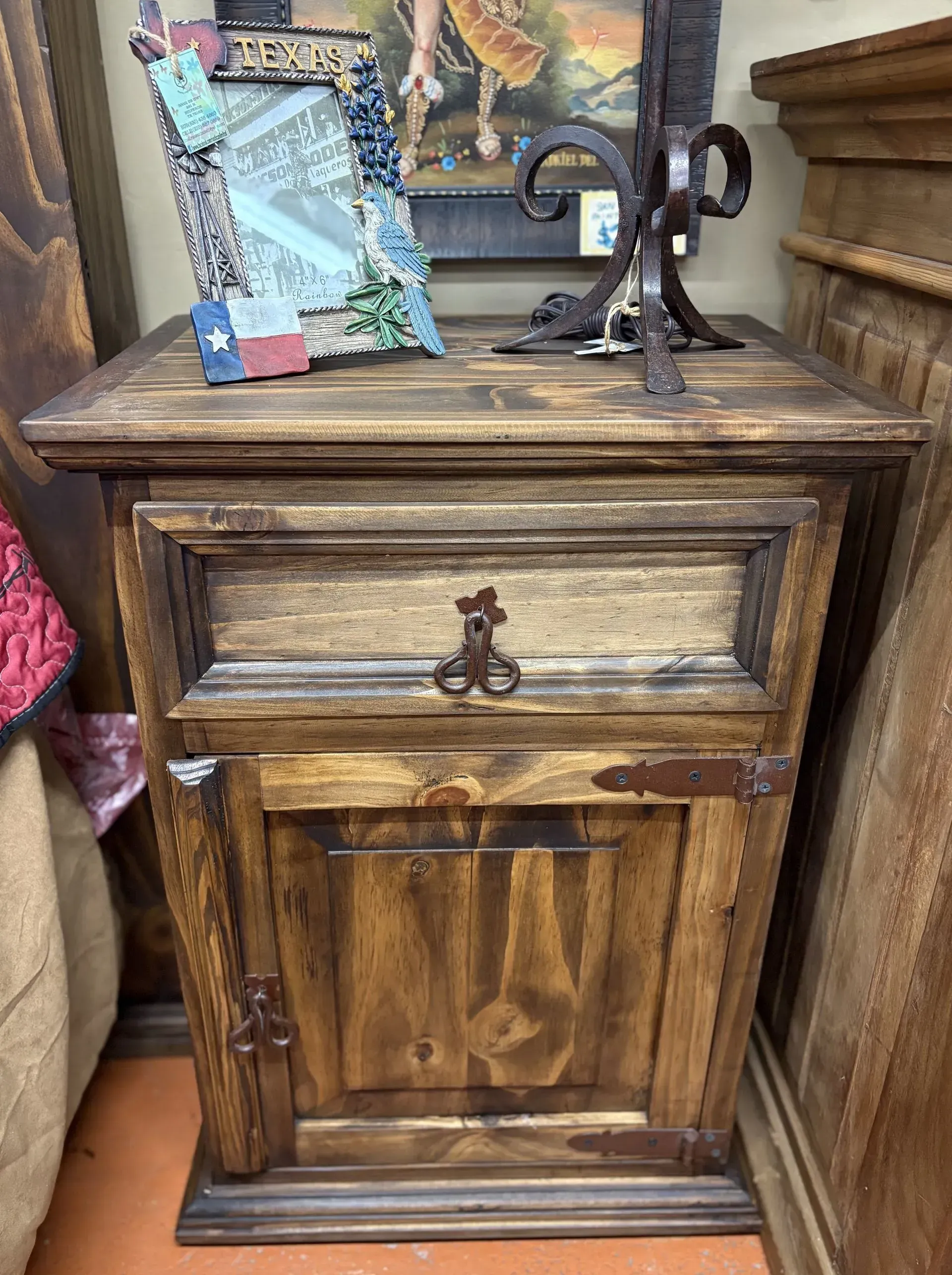 Wooden nightstand with drawer and cabinet, decorated with rustic hardware. The nightstand sits in a room with other wooden furniture.