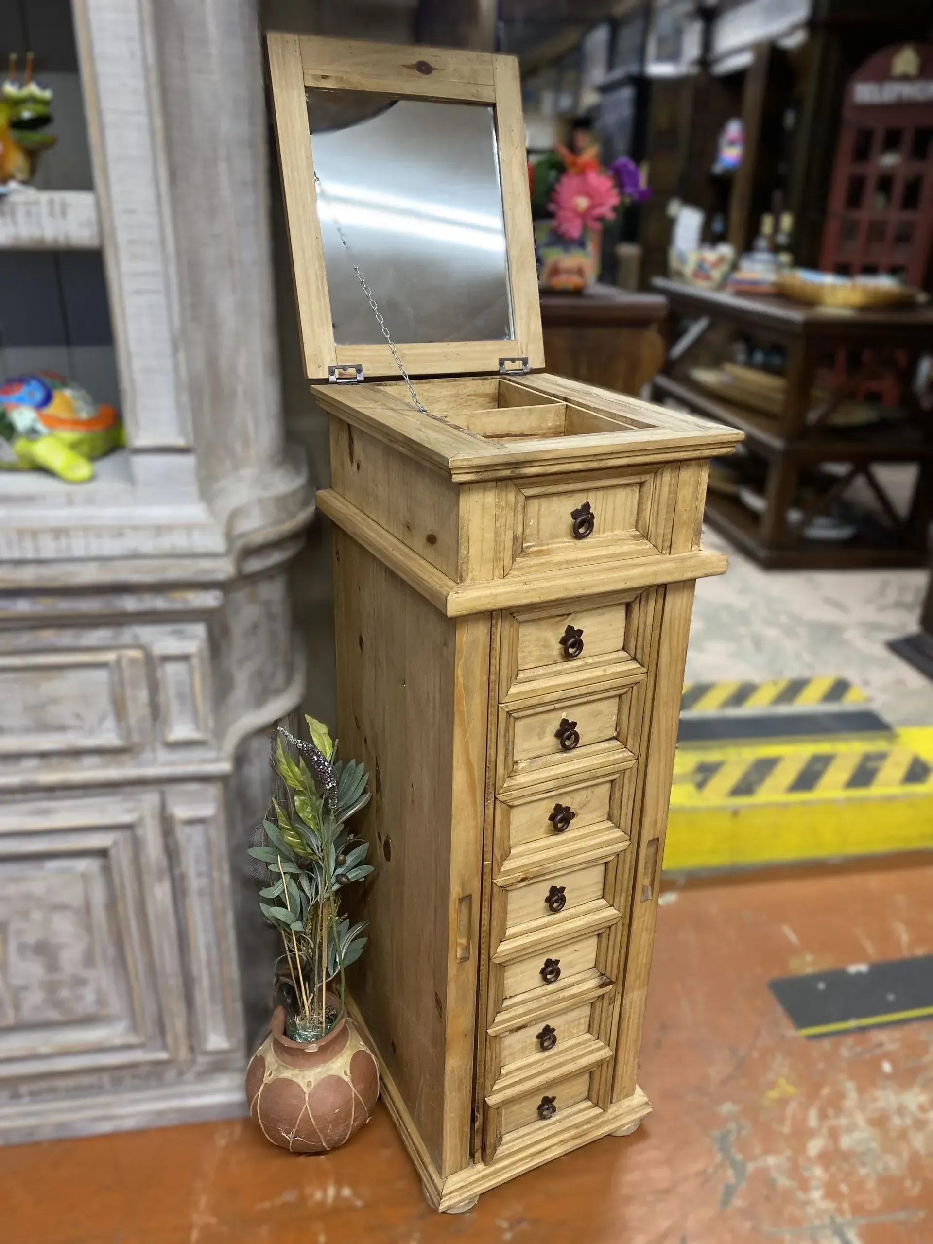 Tall, wooden jewelry armoire with multiple drawers and a built-in mirror, in a shop setting.