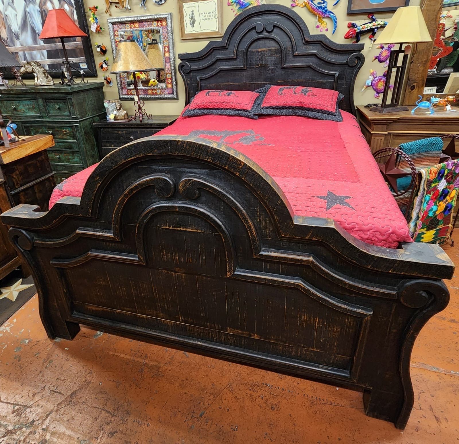 Black wooden bed with ornate headboard and footboard, covered with a red quilt and pillows. The bed is in a cluttered room with other furniture and decor.