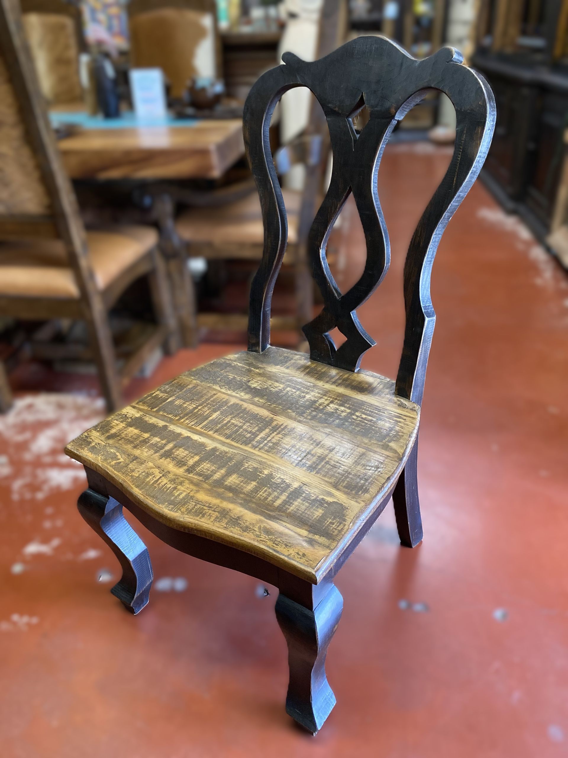 Wooden chair with a dark, ornate back and a distressed, light-brown seat. The chair is on a reddish-orange floor.
