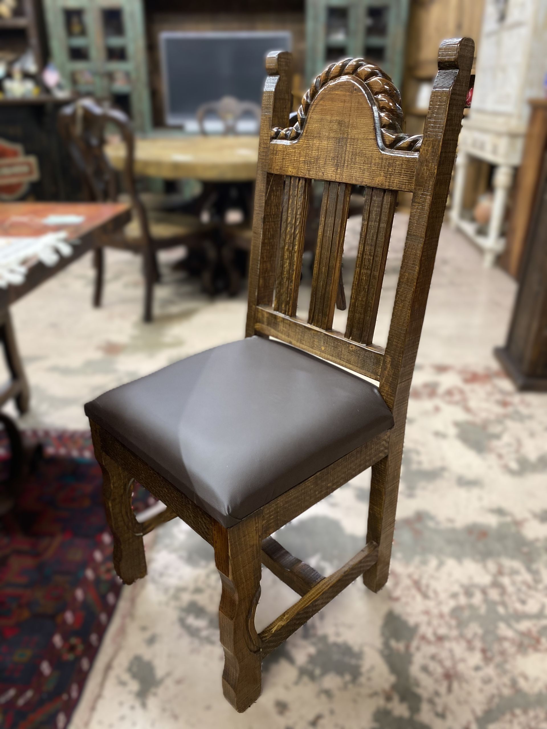 Wooden dining chair with brown leather seat and decorative rope-like carvings on the back. It is set in a cluttered furniture store.