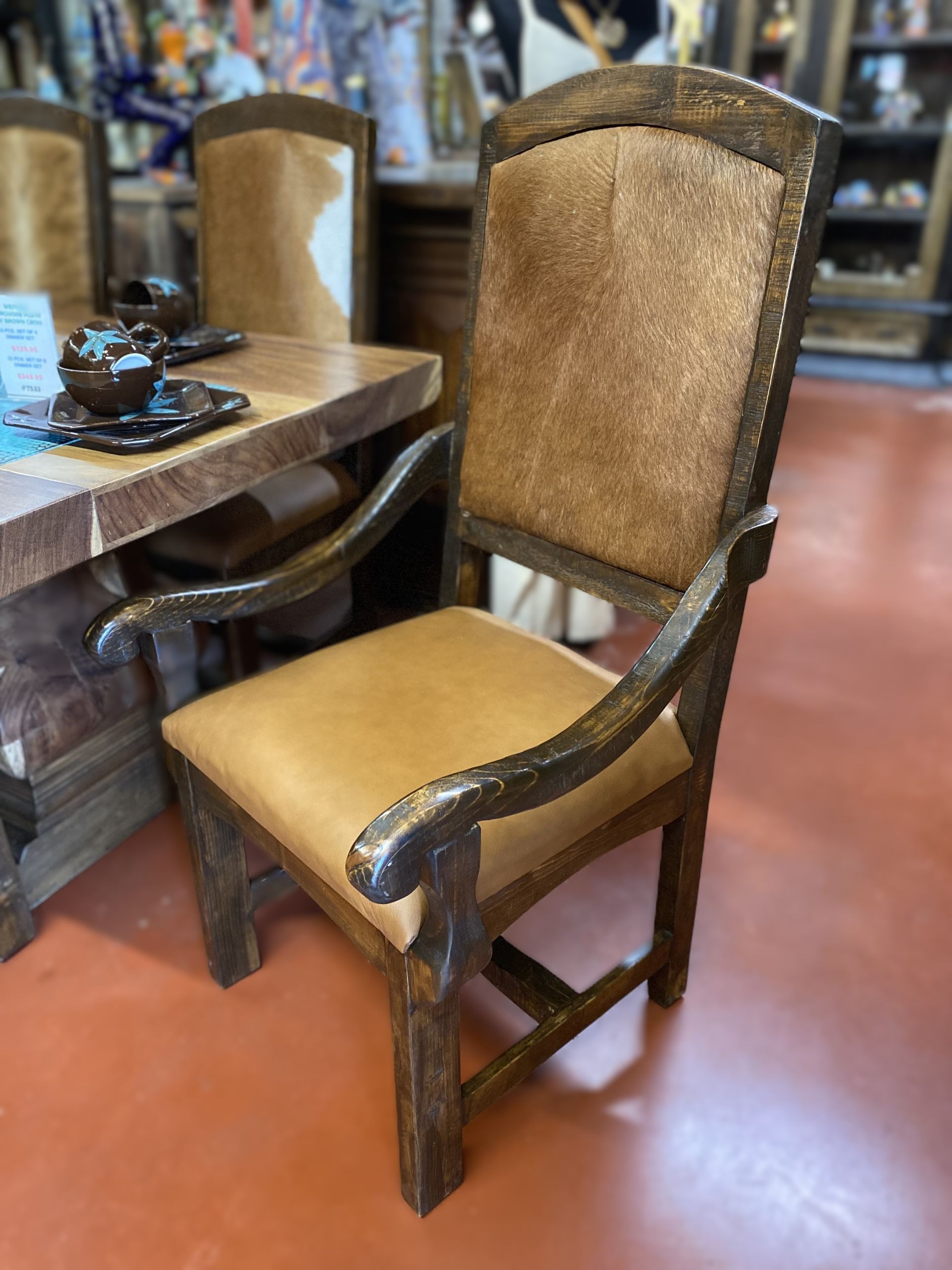 A rustic wooden dining chair with a tan leather seat and a textured back. It is in a showroom with other furniture.