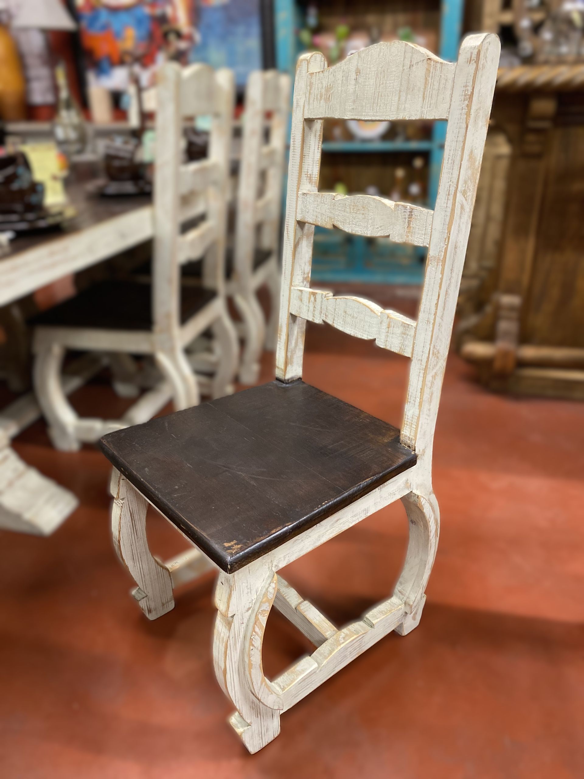 White wooden chair with a dark brown seat, set against a blurred background.  Other chairs and a dining table are visible.