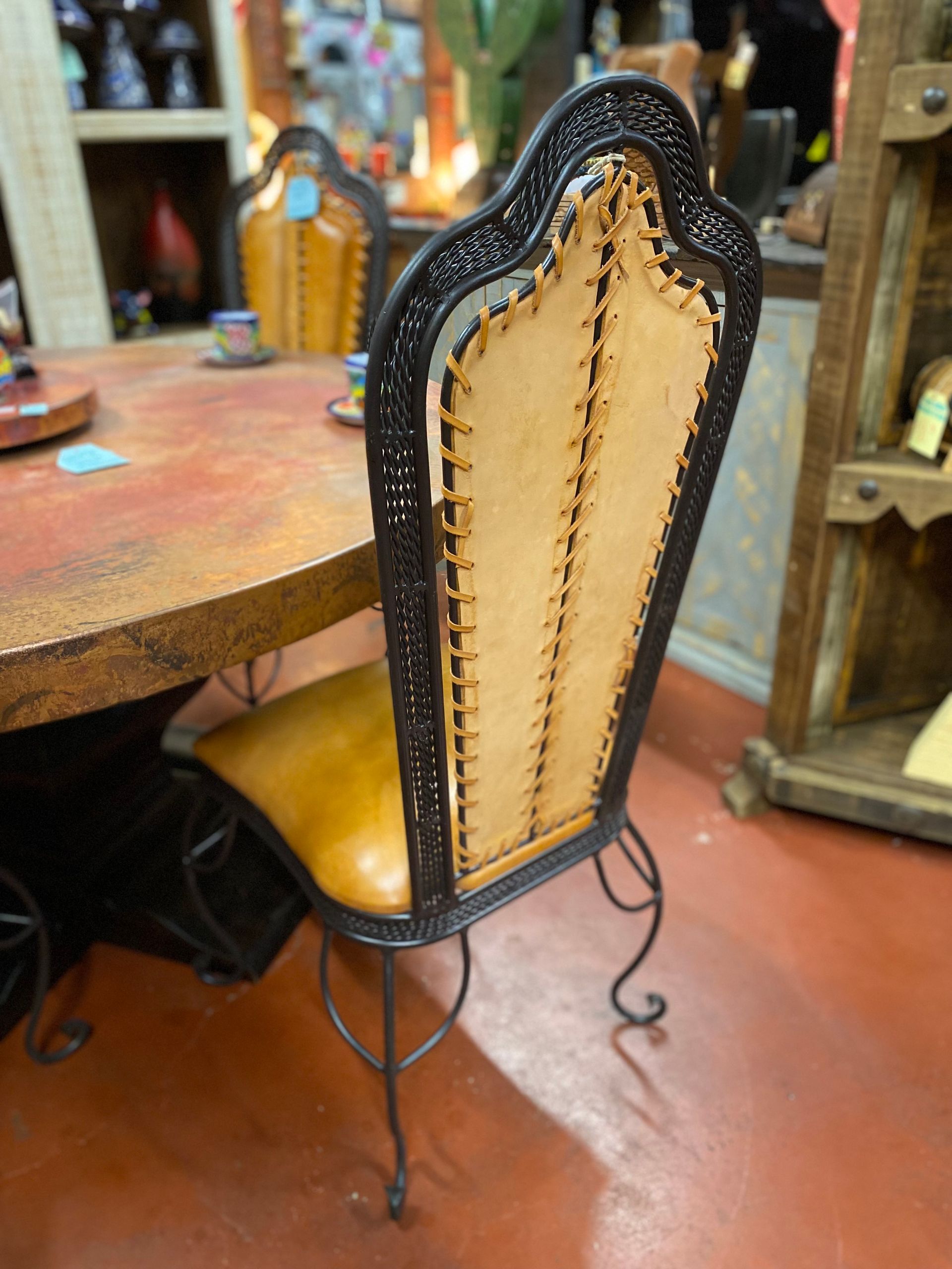 A handcrafted leather and iron chair sits at a rustic table. The chair back is tan leather laced within a dark frame.