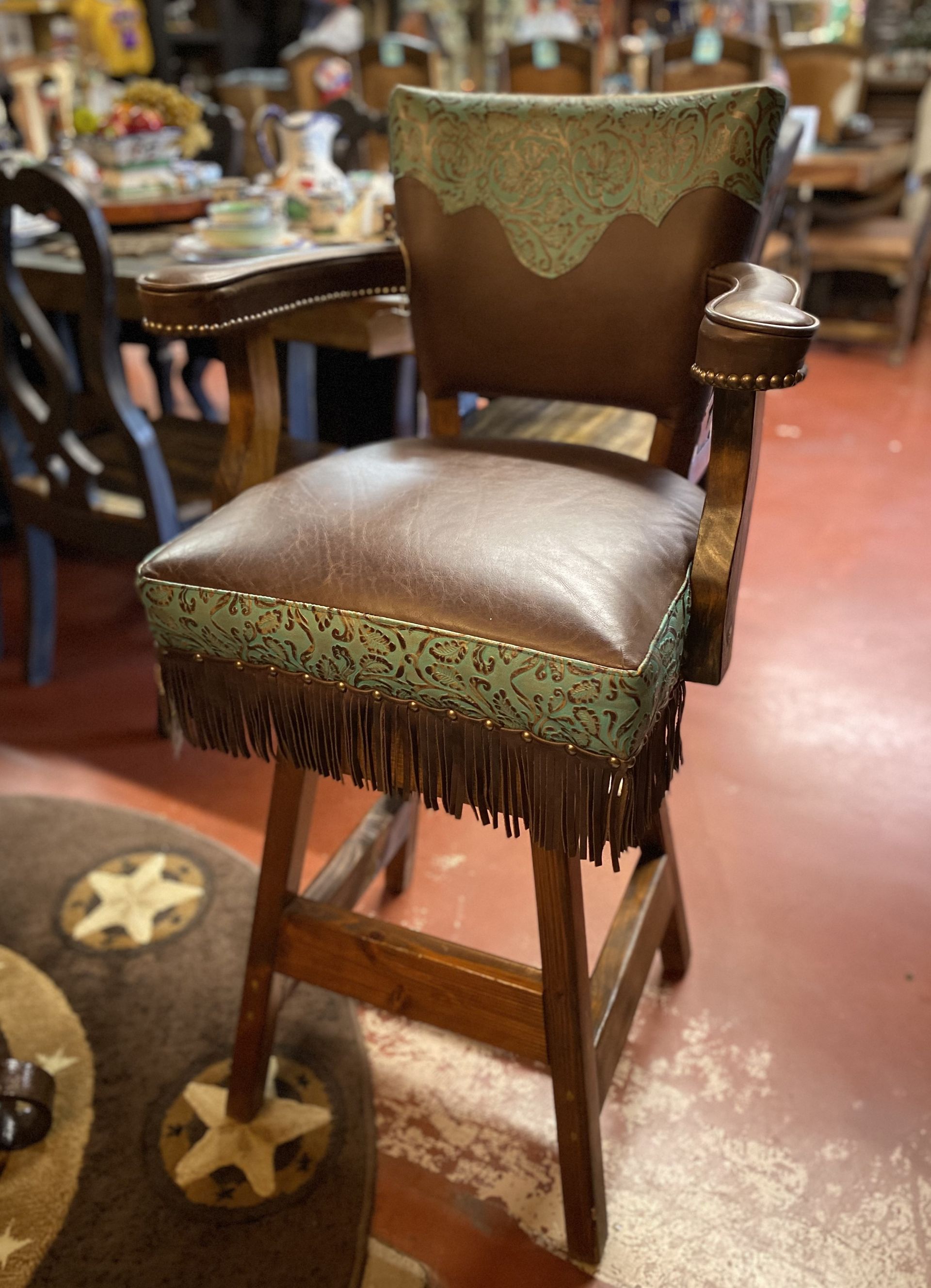 Brown leather and wood bar stool with armrests and turquoise accents; fringed detailing. In a store setting.